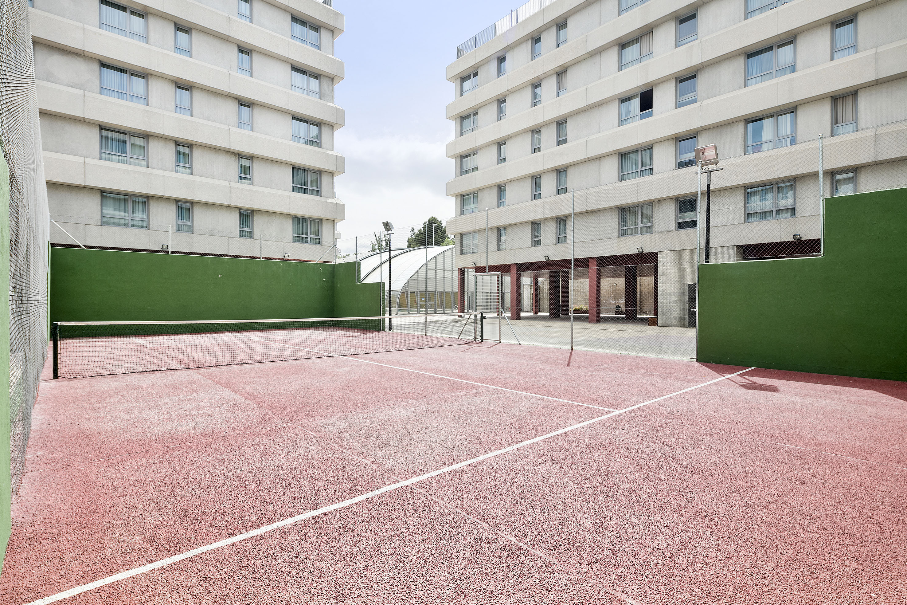 Red padel court with green walls, flanked by modern apartment buildings.