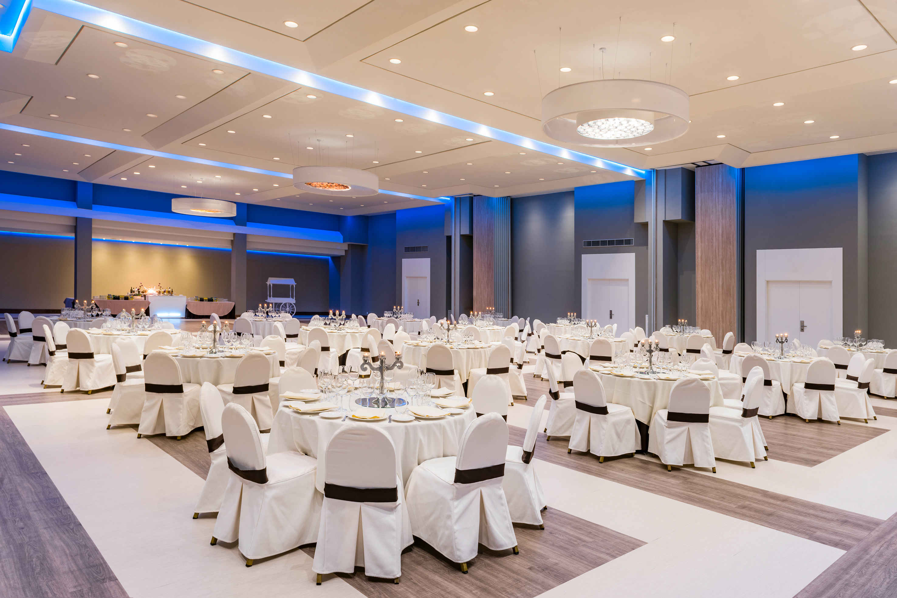 Elegant ballroom with white tables, chairs, modern chandeliers, and blue lighting.