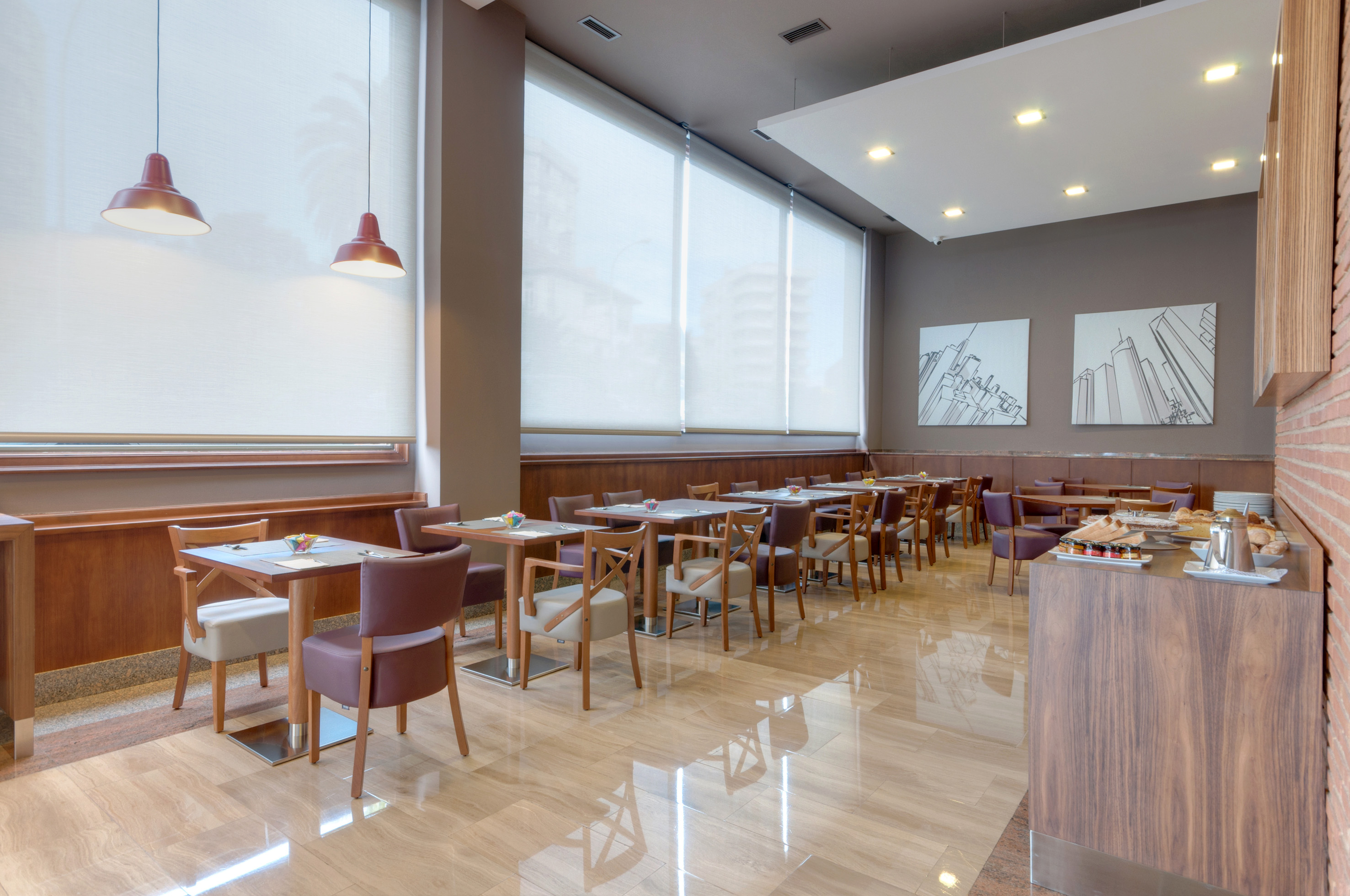 Hotel breakfast room with modern tables, chairs, city art, and buffet.