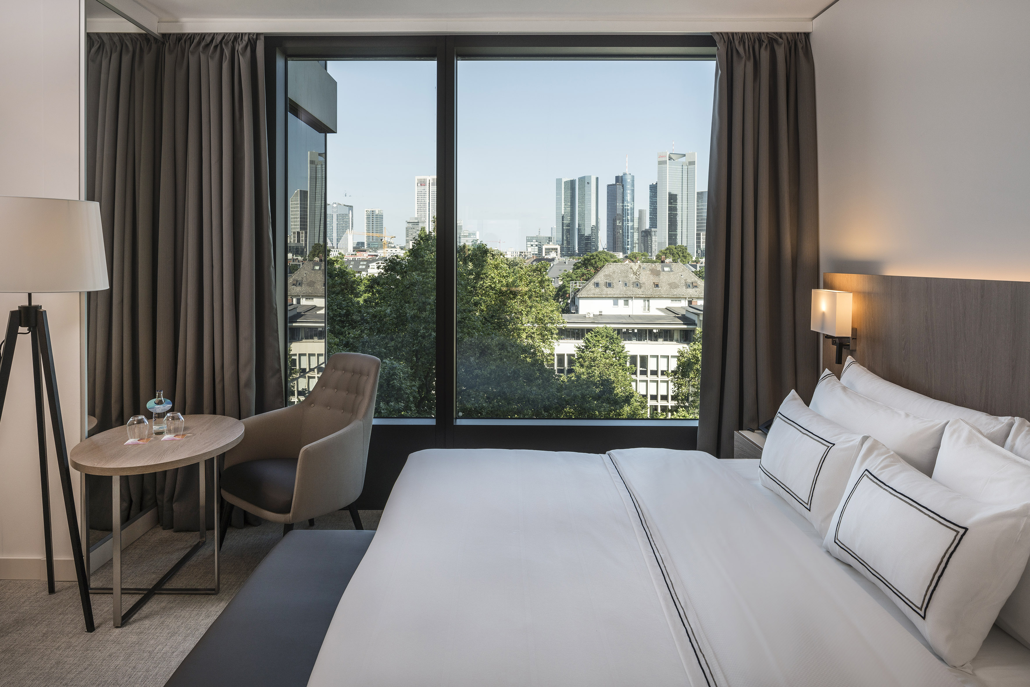 Modern hotel room with king bed, city skyline view, and armchair by window.