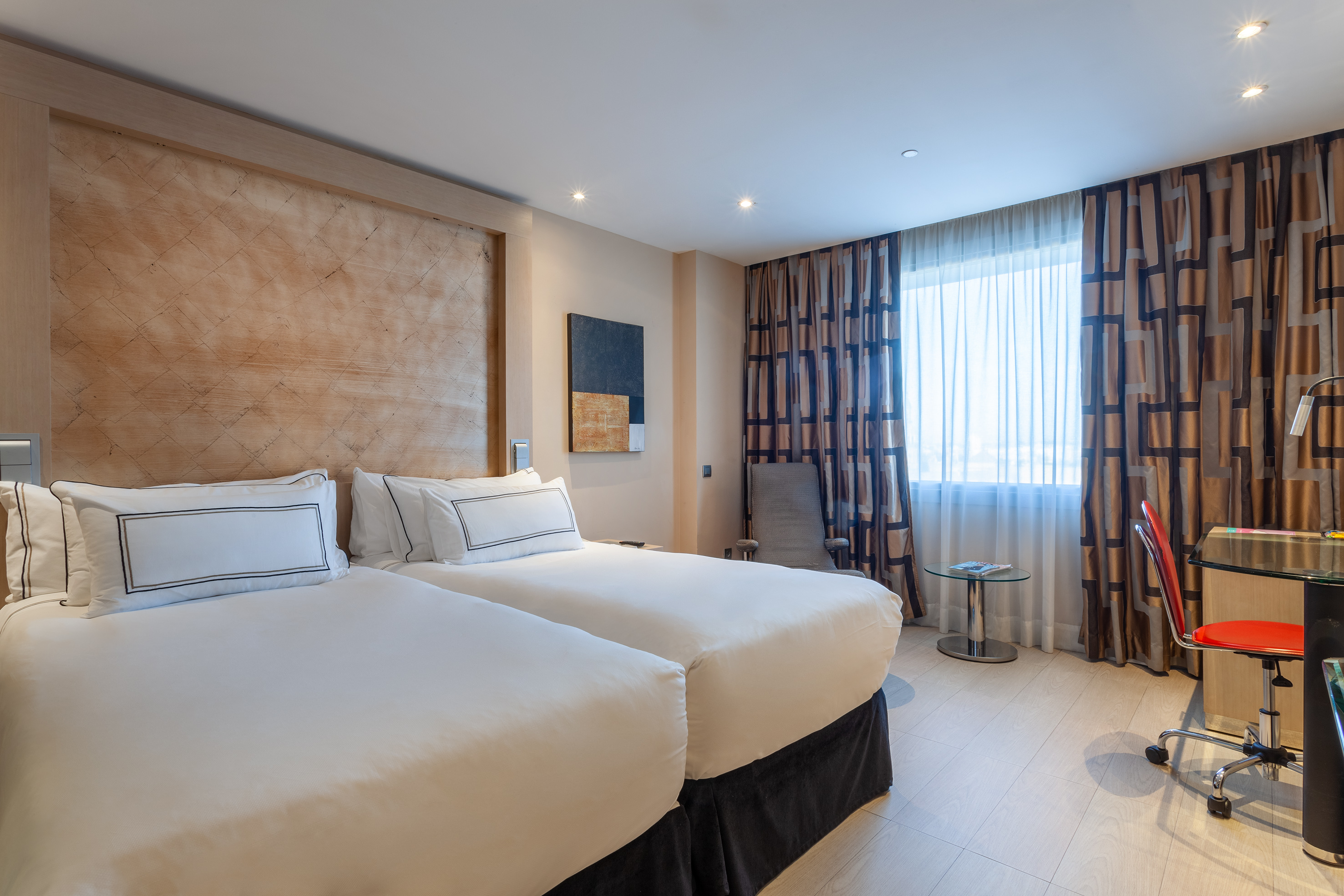 Modern hotel room with two twin beds, patterned curtains, and a desk.
