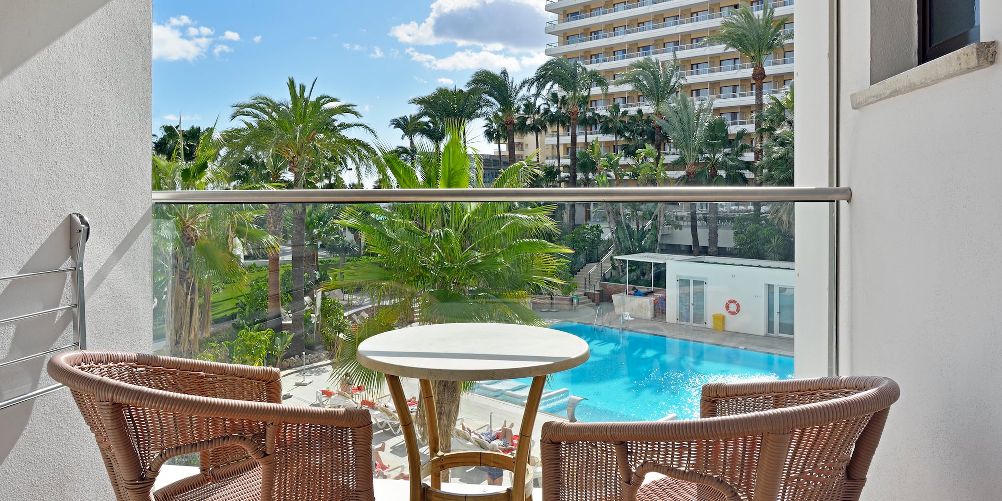 Balcony with rattan furniture, hotel pool, palm trees, and sunny resort view.