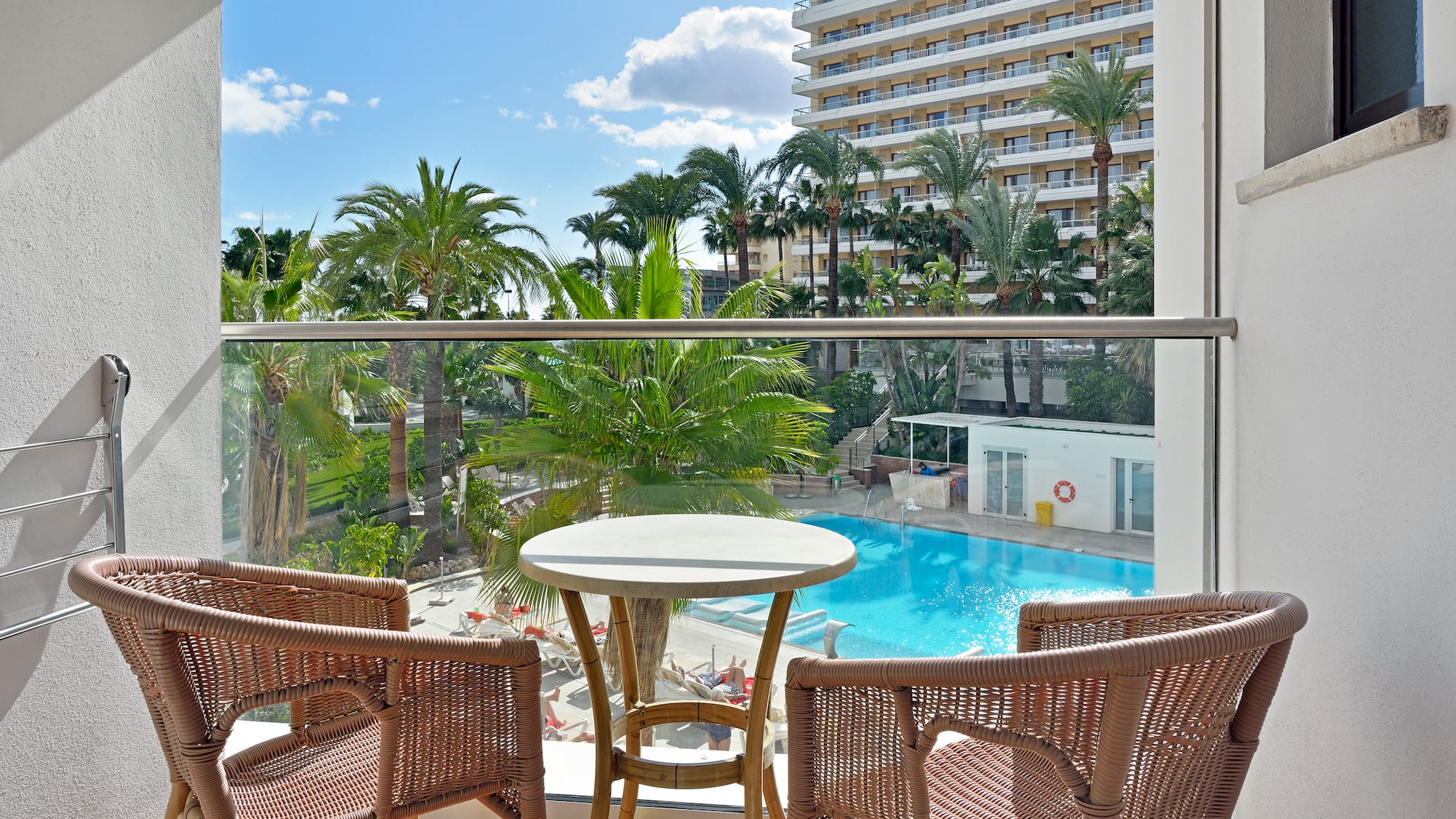 Balcony with rattan furniture, hotel pool, palm trees, and sunny resort view.