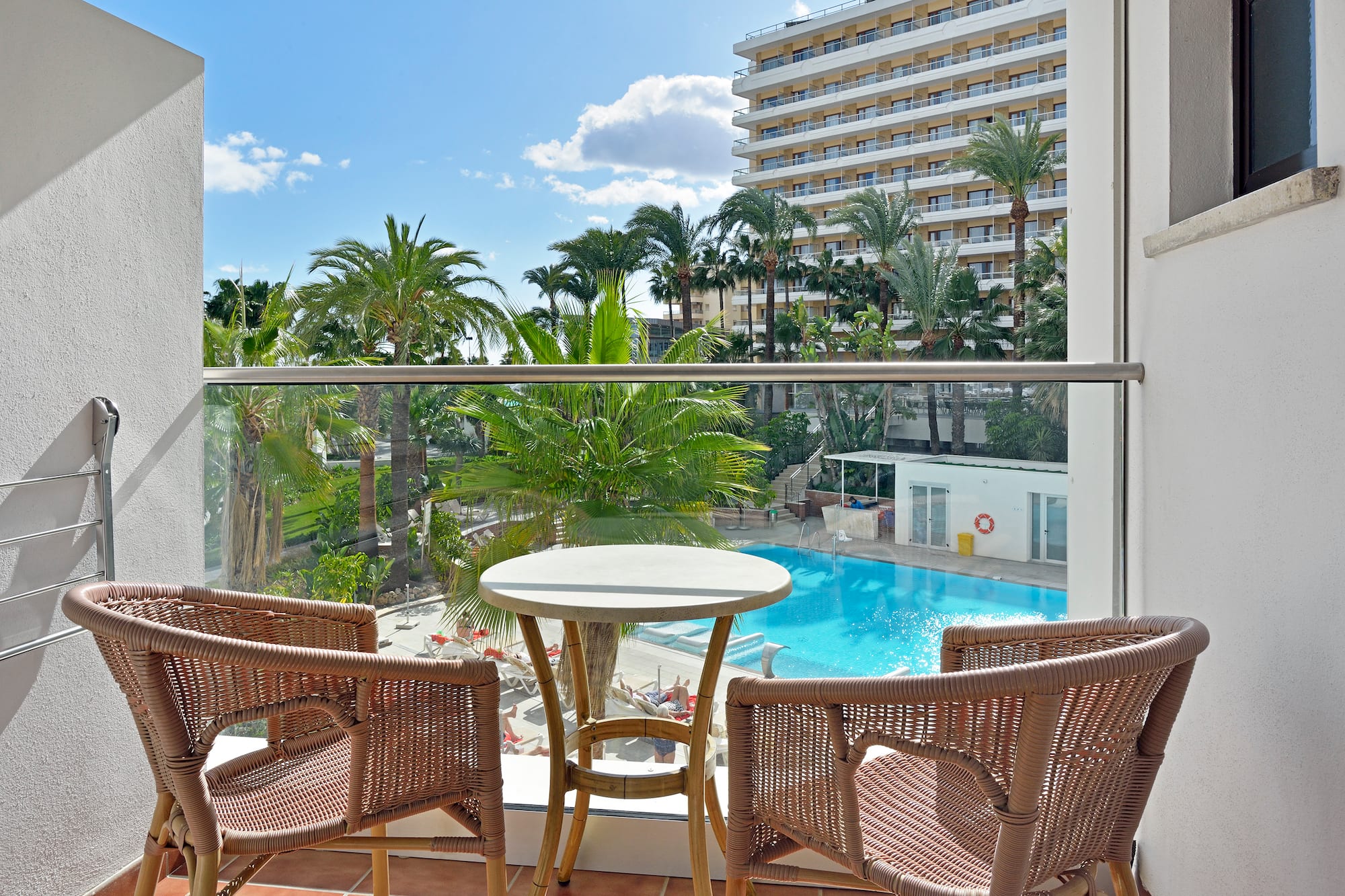 Balcony with rattan furniture, hotel pool, palm trees, and sunny resort view.