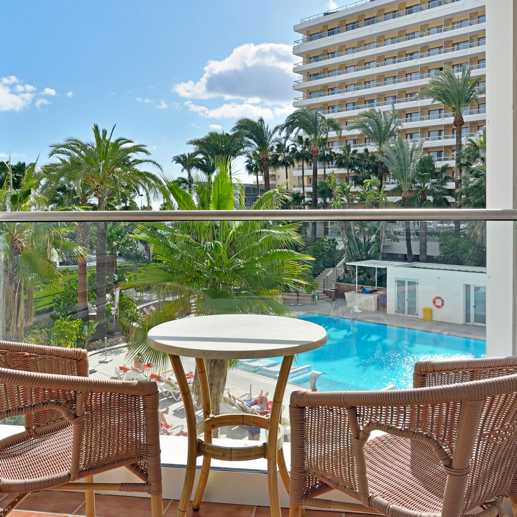 Balcony with rattan furniture, hotel pool, palm trees, and sunny resort view.