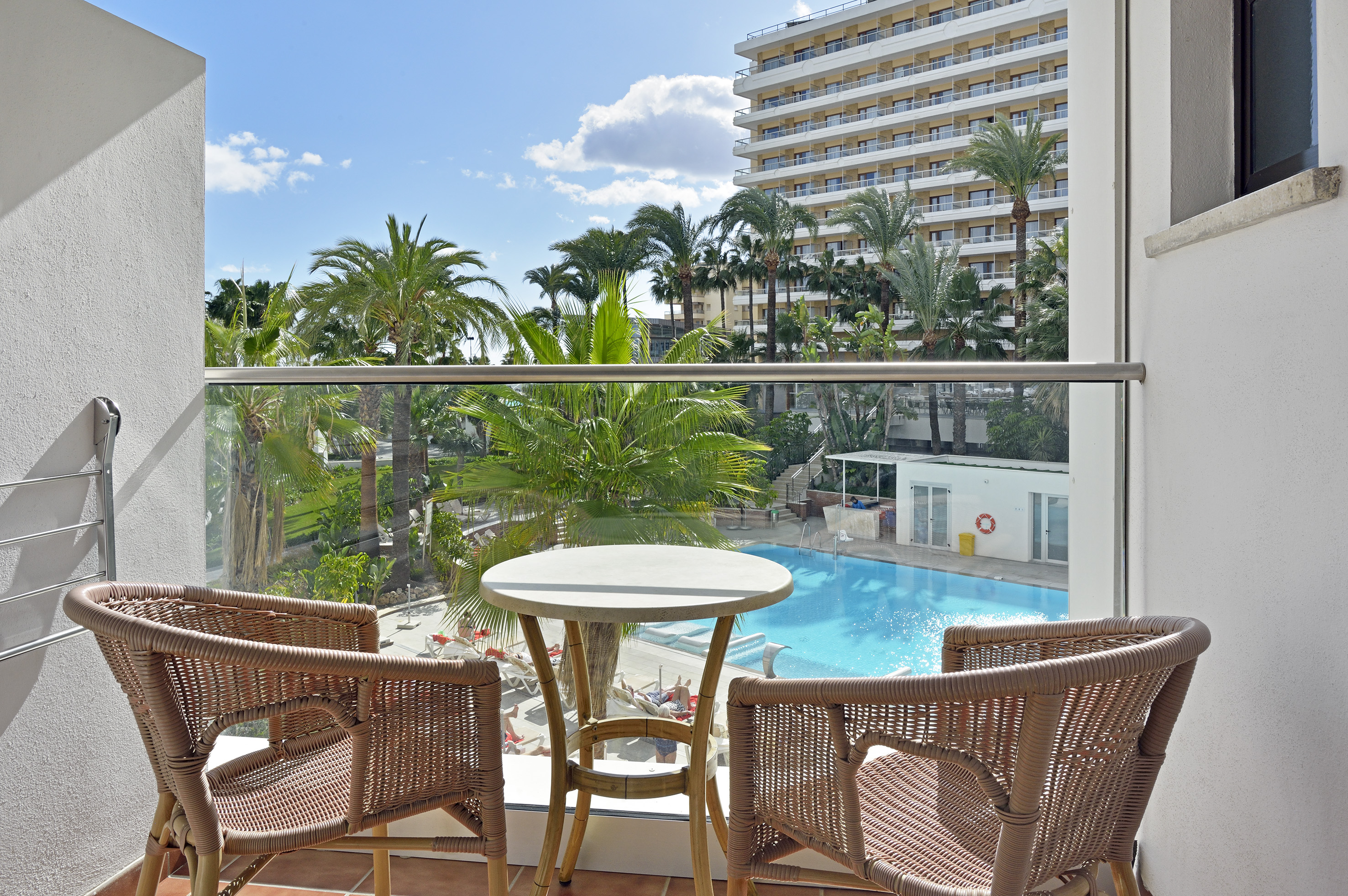 Balcony with rattan furniture, hotel pool, palm trees, and sunny resort view.