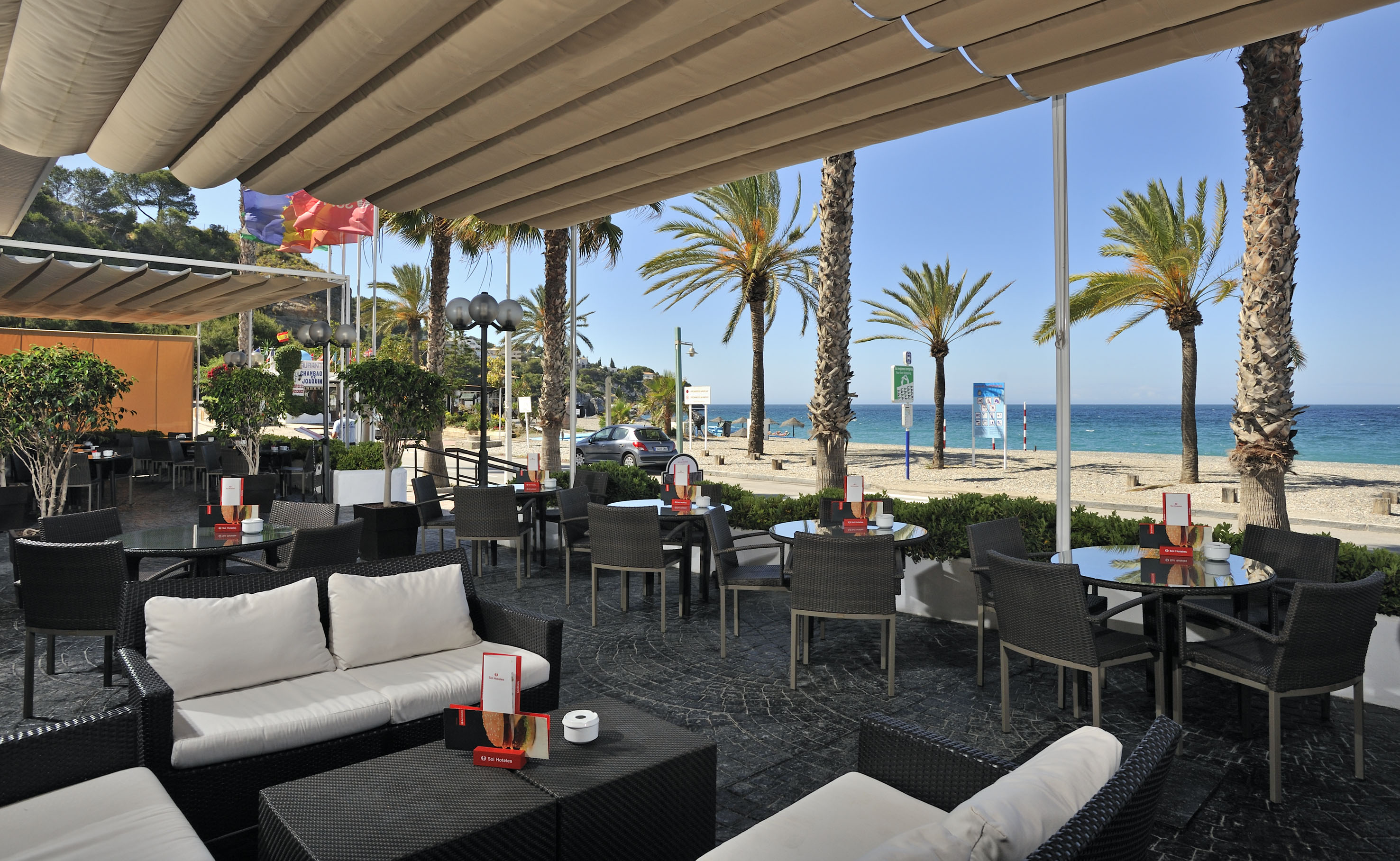 Beachfront outdoor patio with cafe seating, palm trees, and clear blue sea.