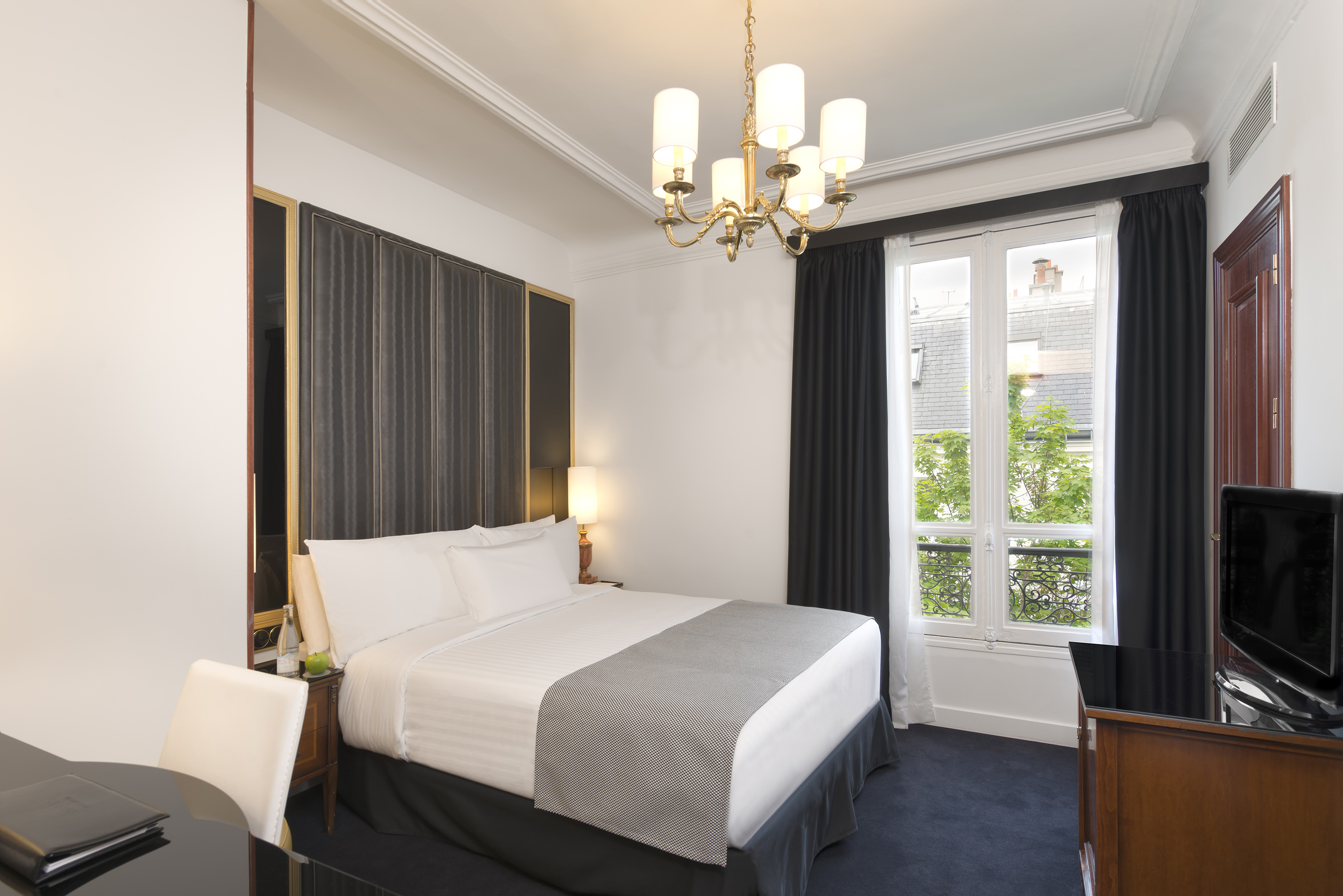 Elegant hotel room with white bed, golden chandelier, desk, and large window.