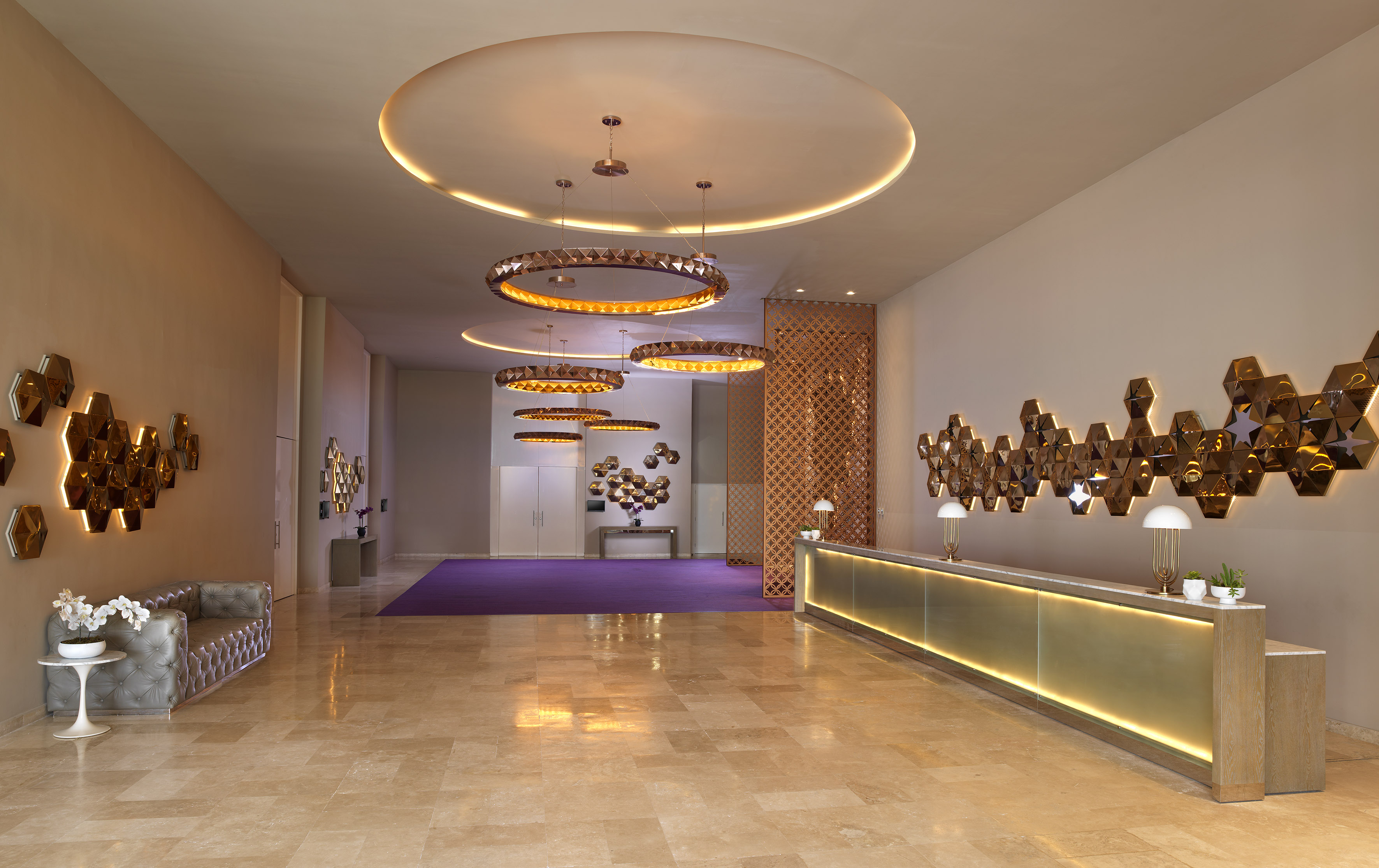 Modern hotel reception area with golden desk, hexagonal wall art, circular lights.