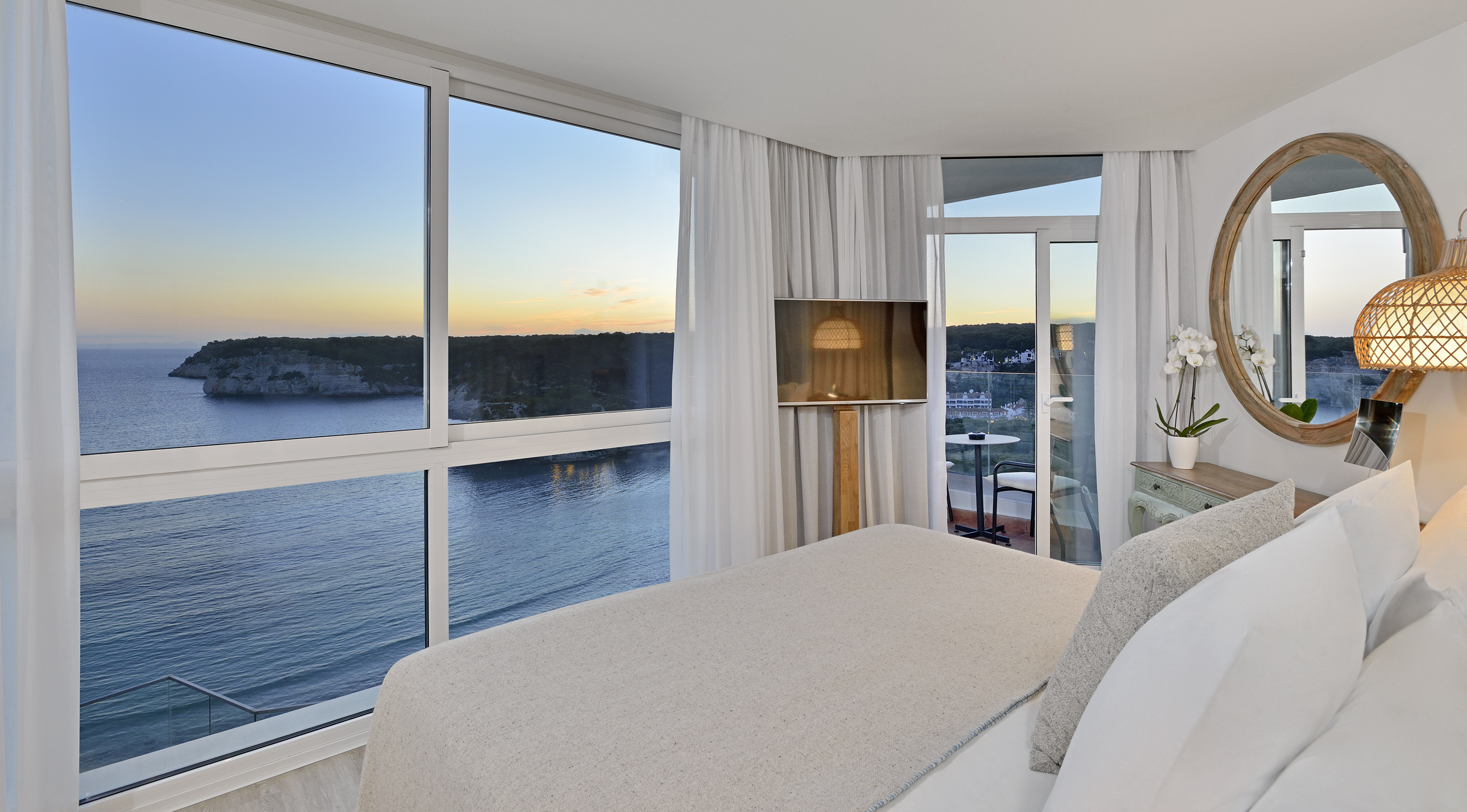 Modern hotel bedroom with panoramic ocean view at sunset, private balcony.