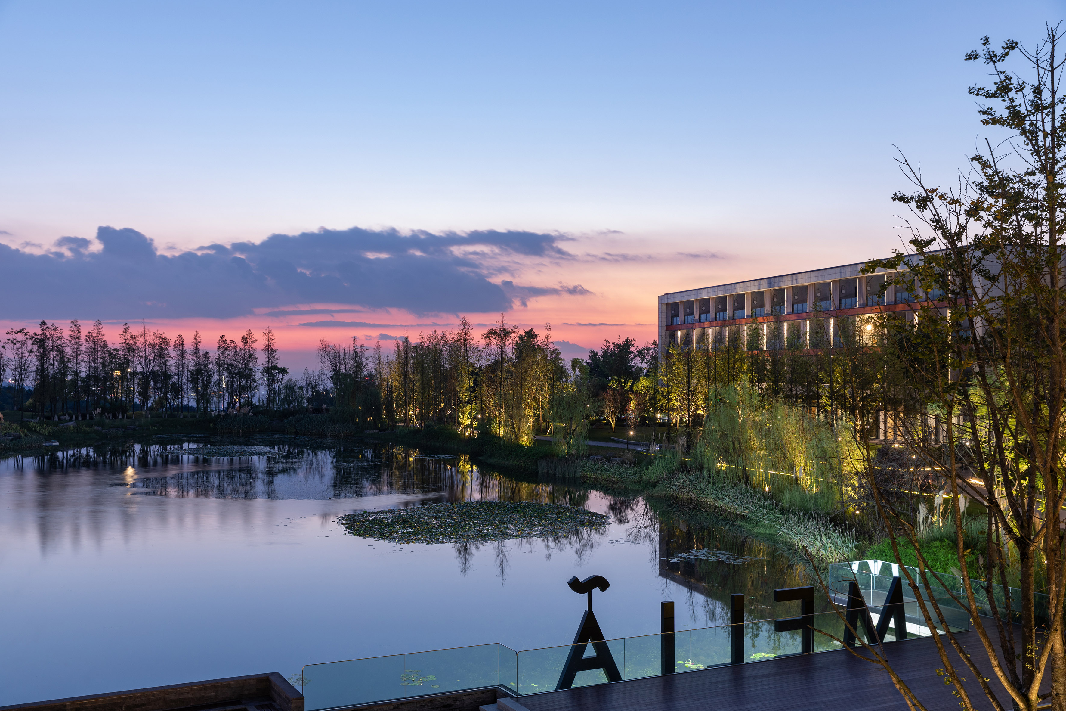 Modern hotel at sunset overlooking a tranquil lake with illuminated trees and unique art.