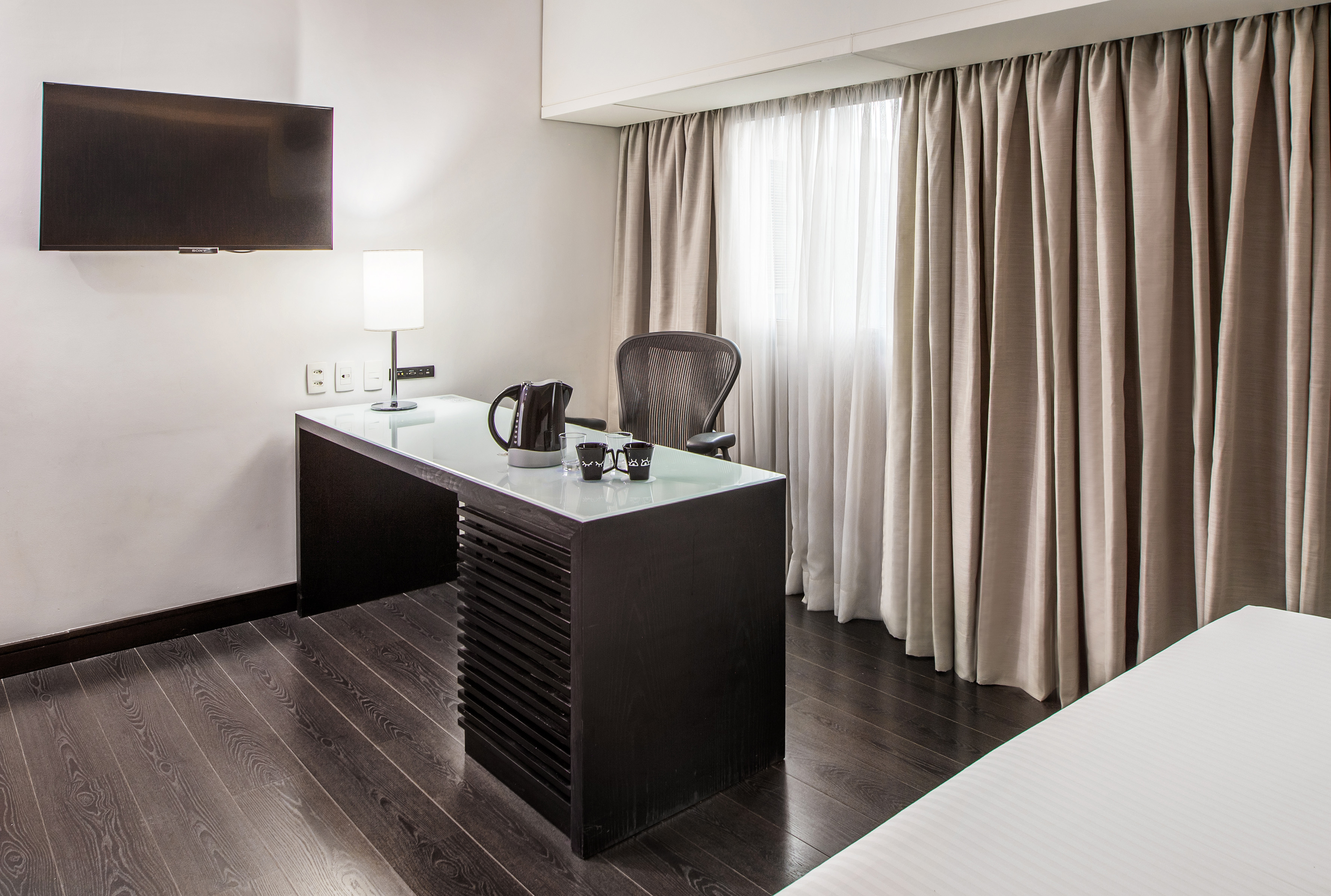 Modern hotel room with work desk, TV, kettle, and beige curtains.