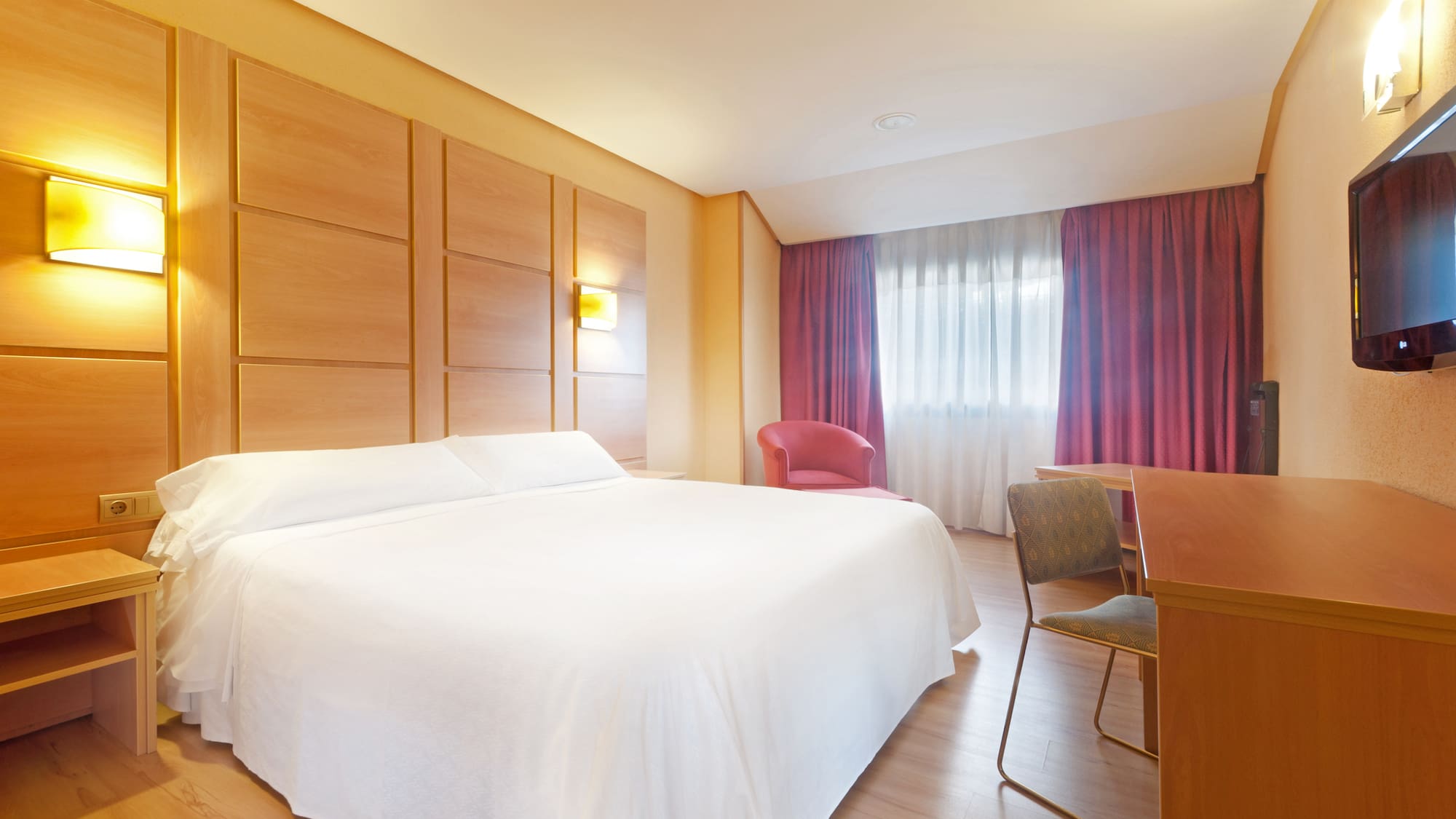Modern hotel bedroom with white bed, wood headboard, red curtains, and work desk.