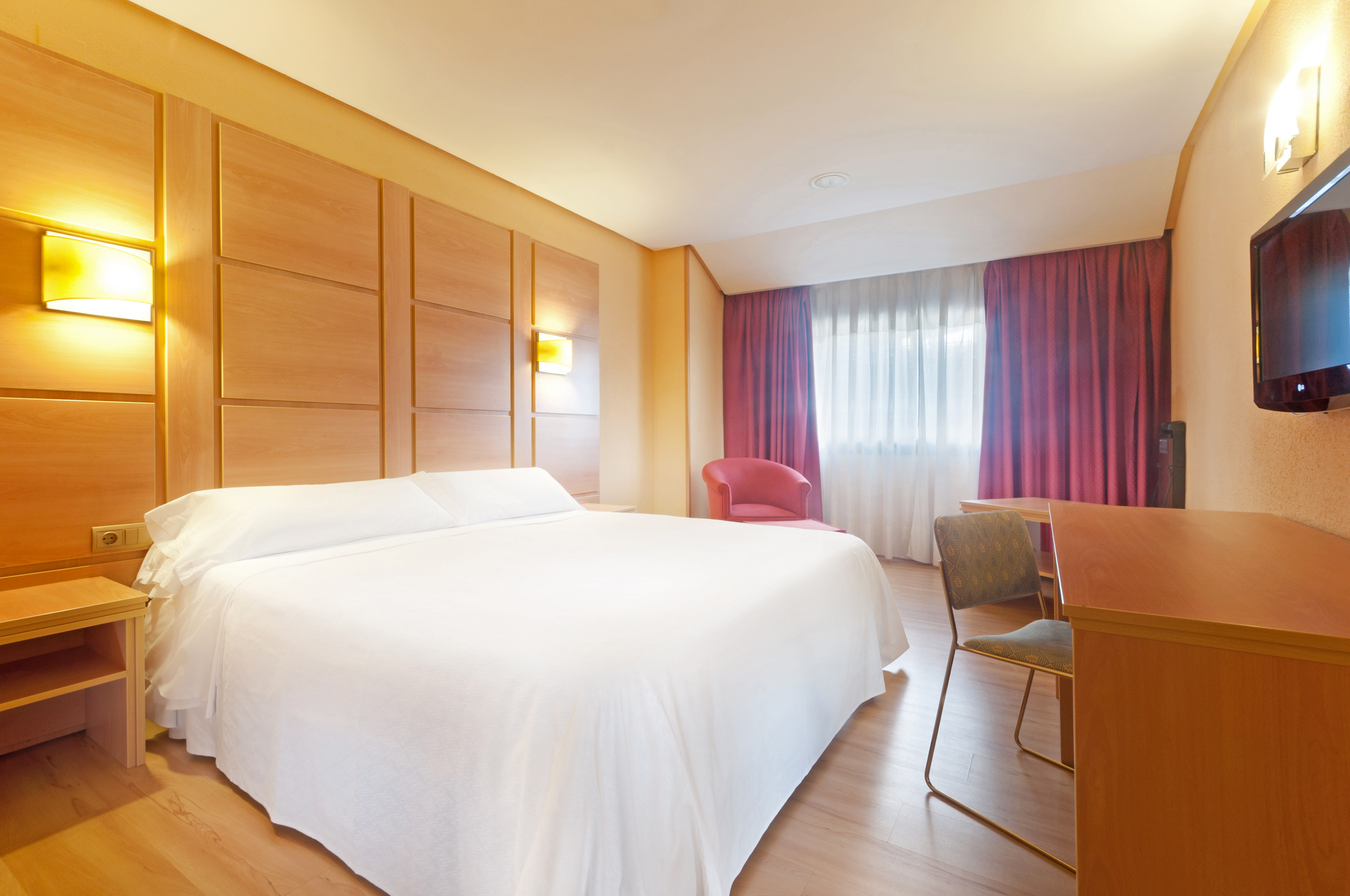 Modern hotel bedroom with white bed, wood headboard, red curtains, and work desk.