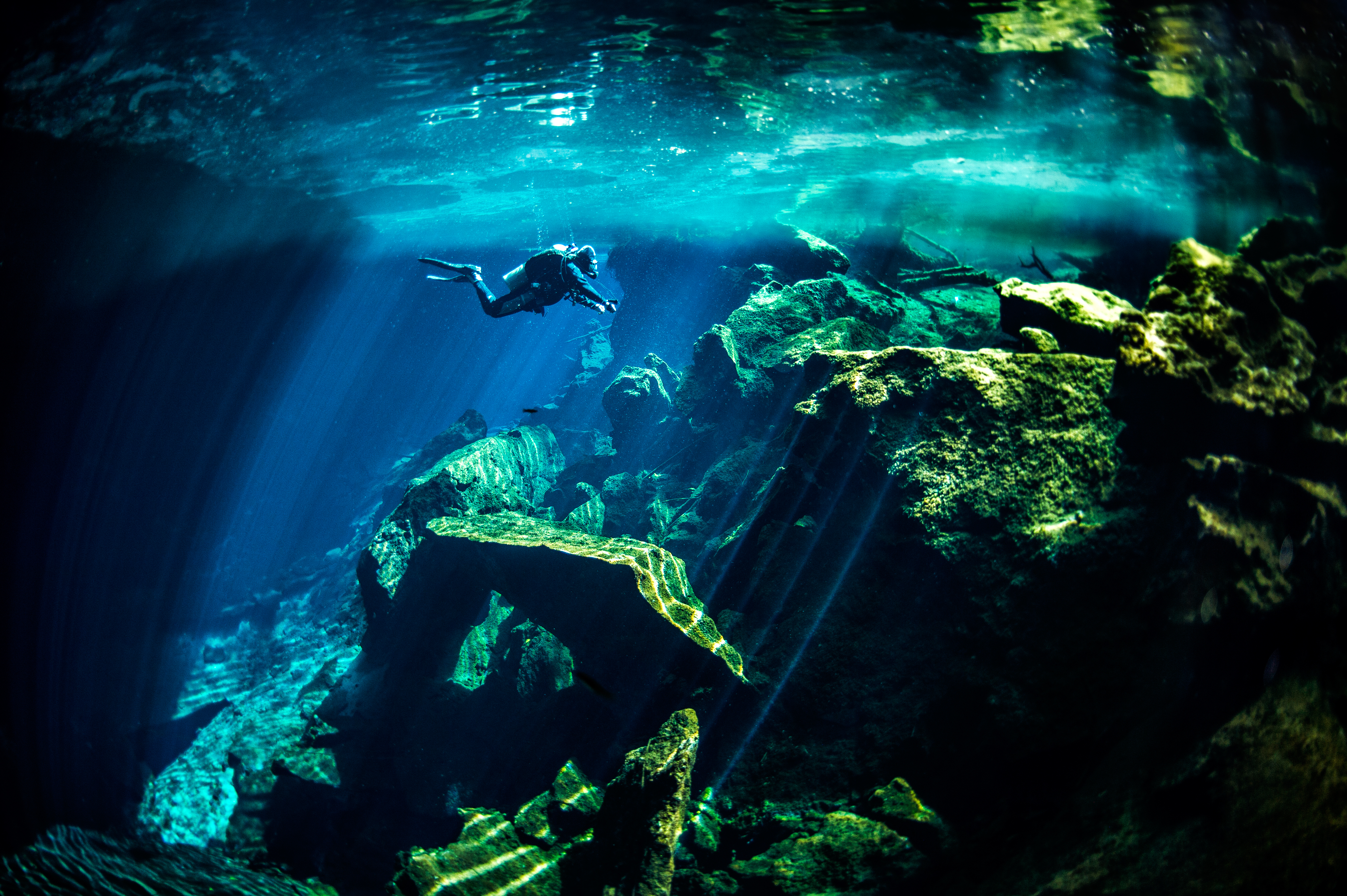 Buceador explorando cenote con rayos de sol entre rocas.