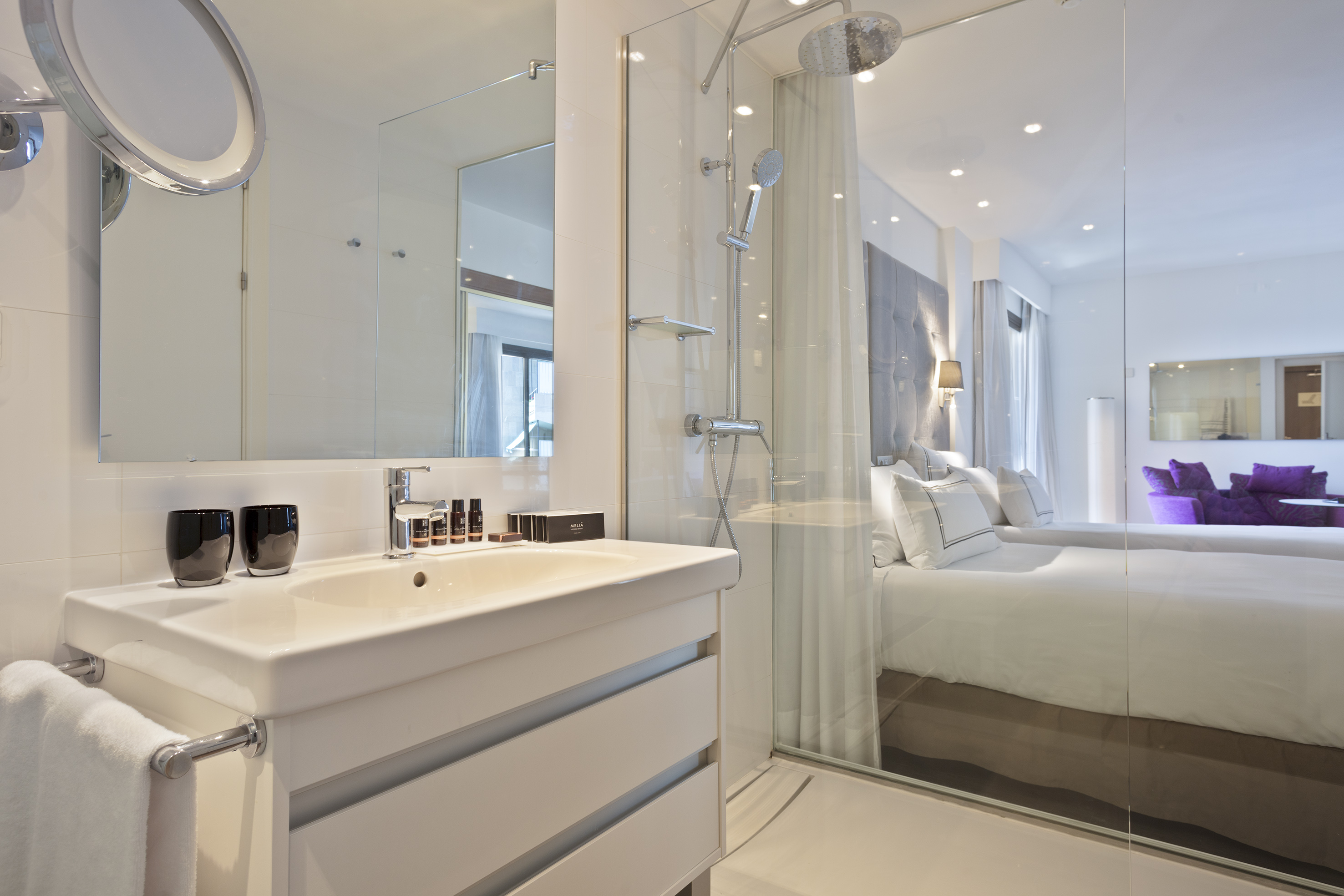 Modern hotel bathroom with sink, glass shower, and bedroom view.
