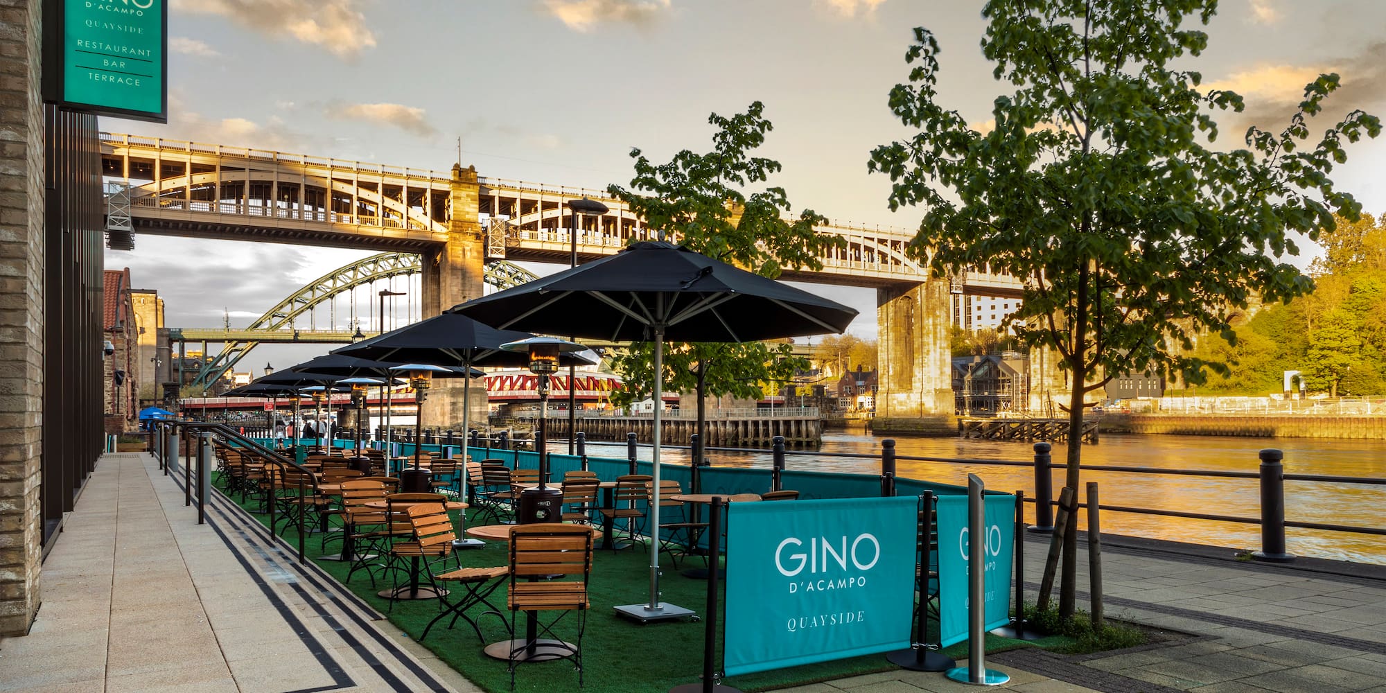 a group of tables and chairs on a grass area with umbrellas and a bridge in the background