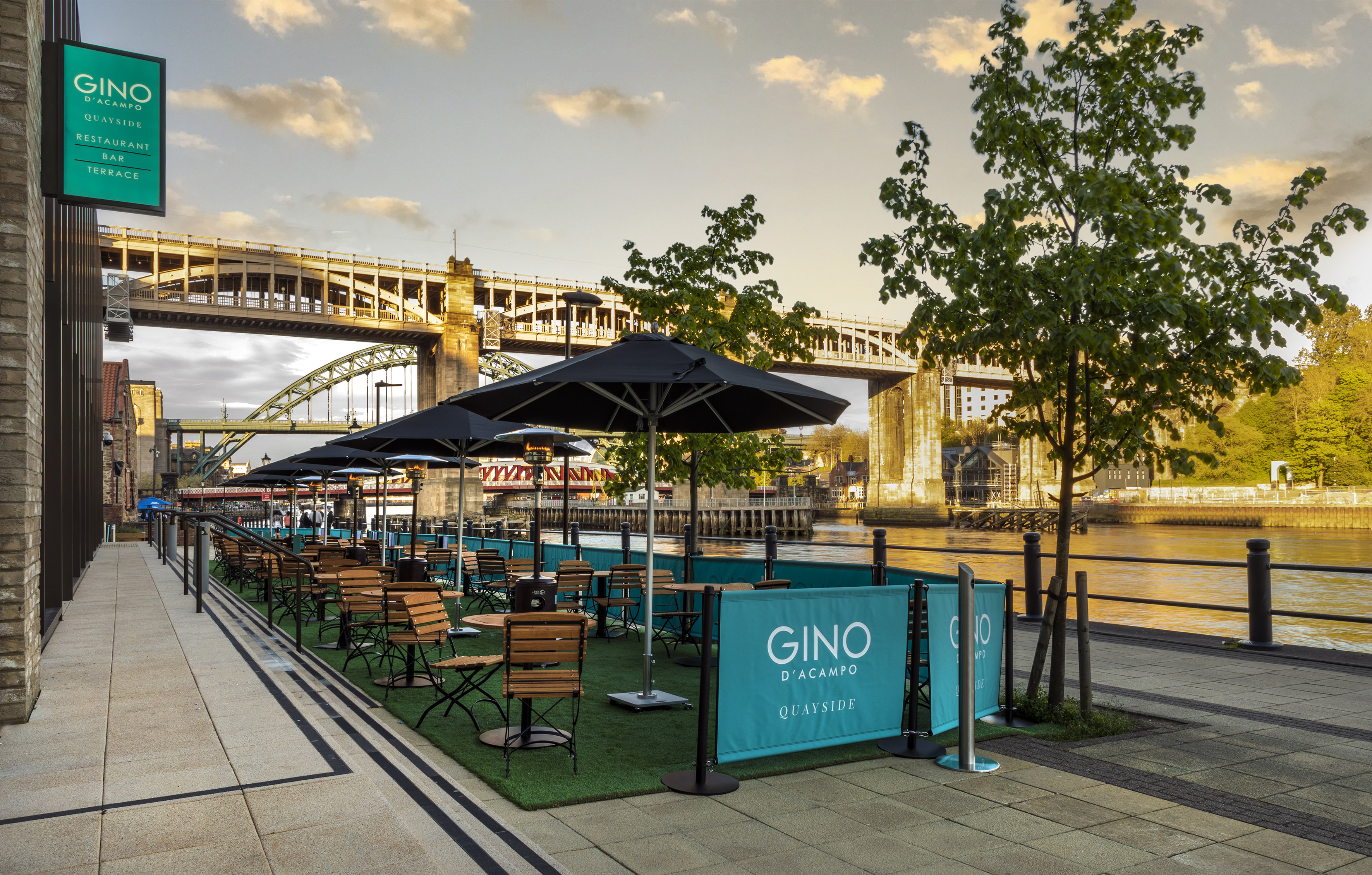 a group of tables and chairs on a grass area with umbrellas and a bridge in the background