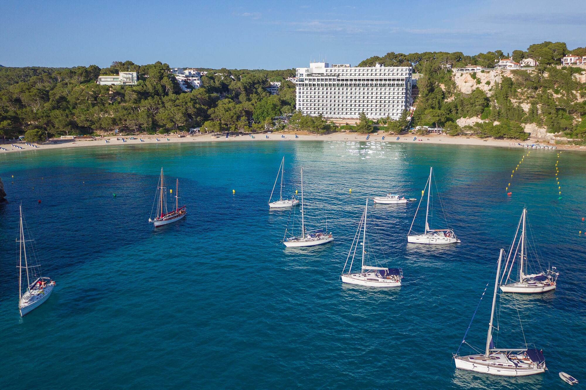 Sailboats in Mediterranean bay with sandy beach and large resort hotel.