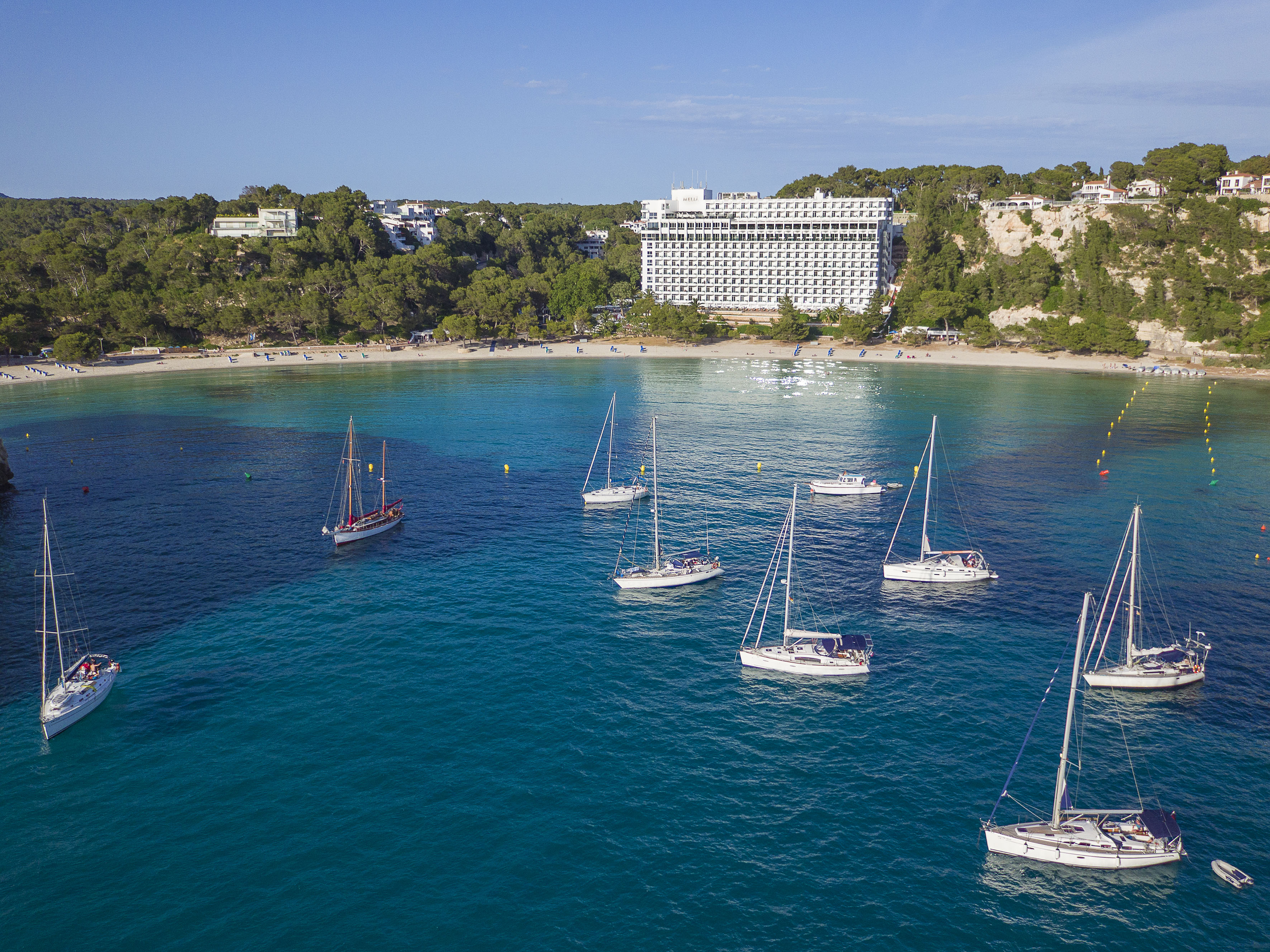 Sailboats in Mediterranean bay with sandy beach and large resort hotel.