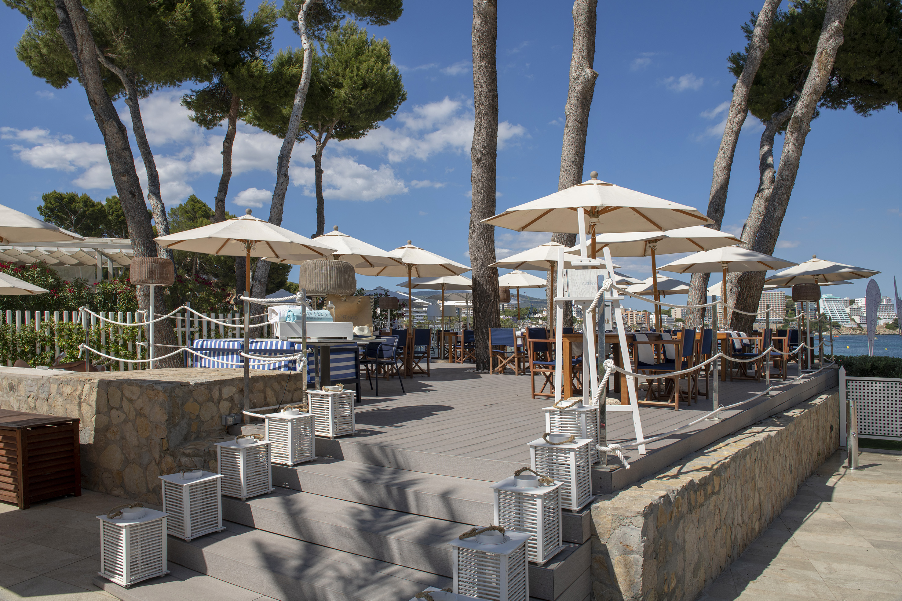 Beach club restaurant terrace with umbrellas, dining tables, white lanterns, and sea view.