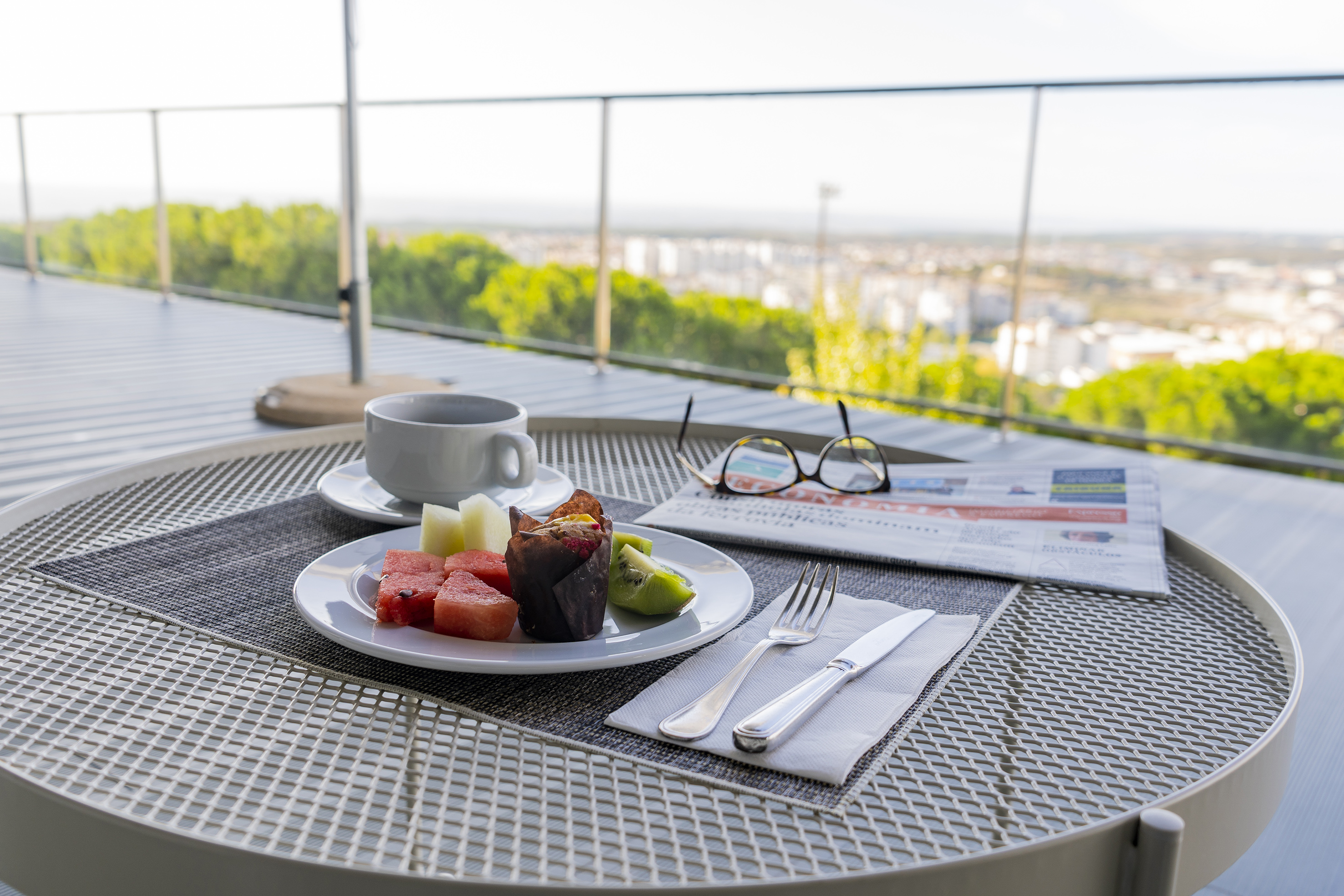 Breakfast on a balcony with coffee, fresh fruit, muffin, newspaper, and city view.