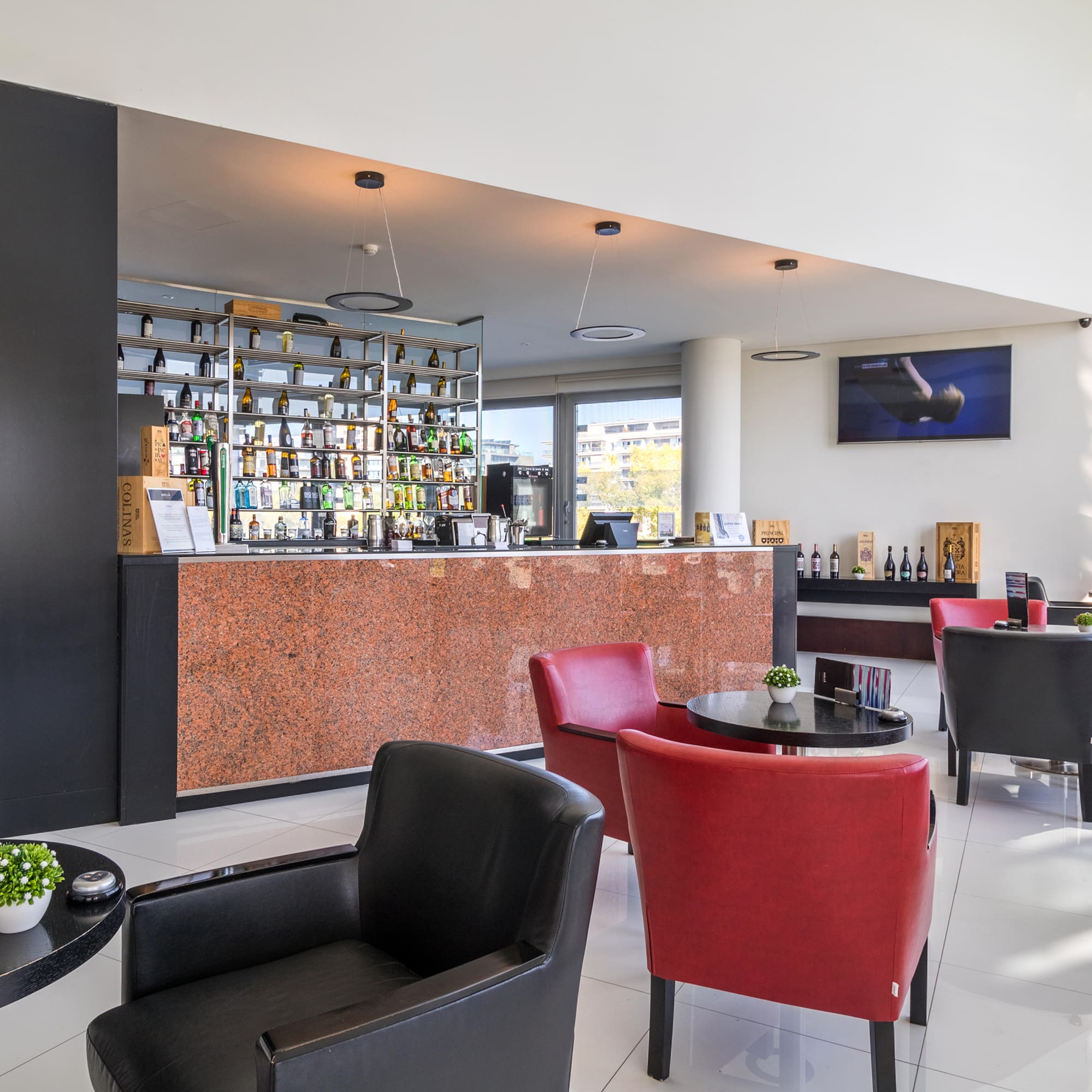 Modern hotel bar and lounge with liquor shelf, granite counter, red & black armchairs.
