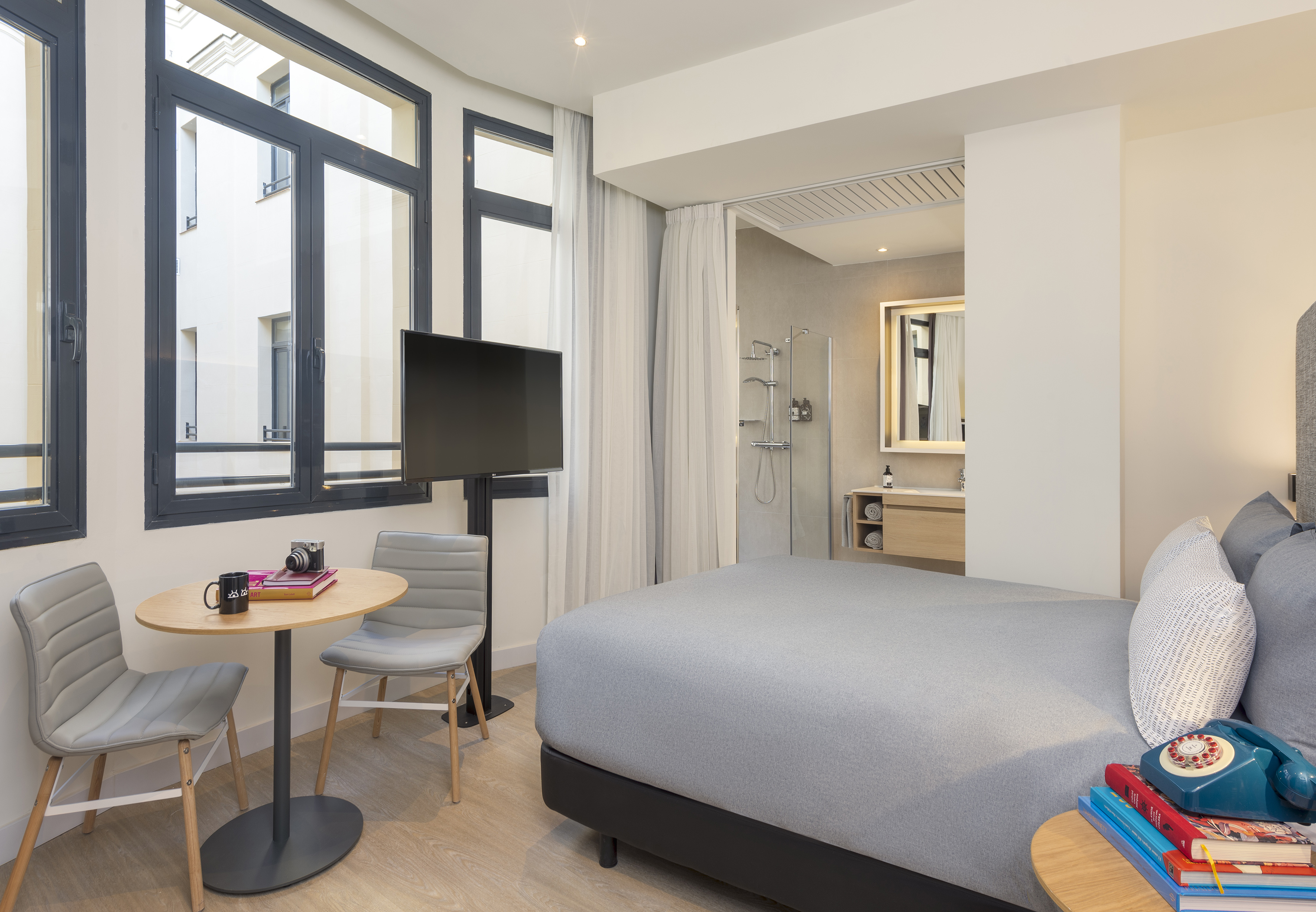Stylish hotel room with grey bed, window, small table, TV, and open shower.