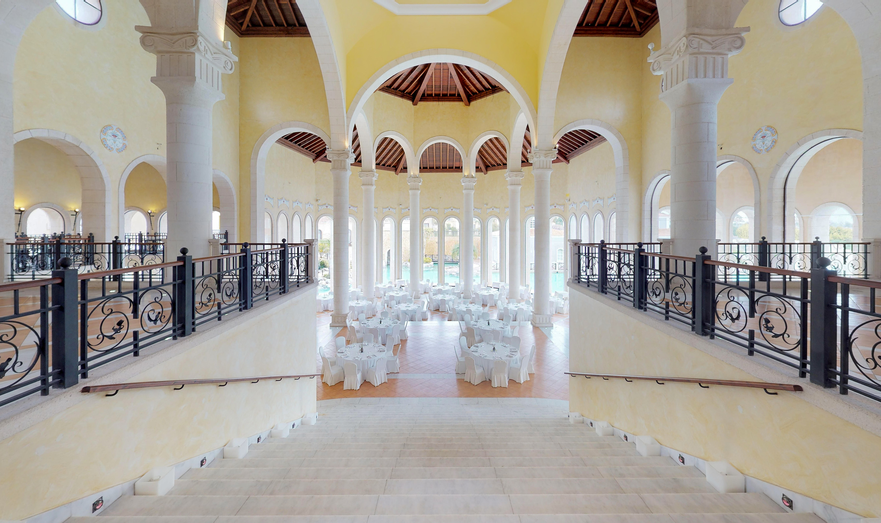 Grand ballroom with white event tables, towering columns, arches, and elegant staircase.
