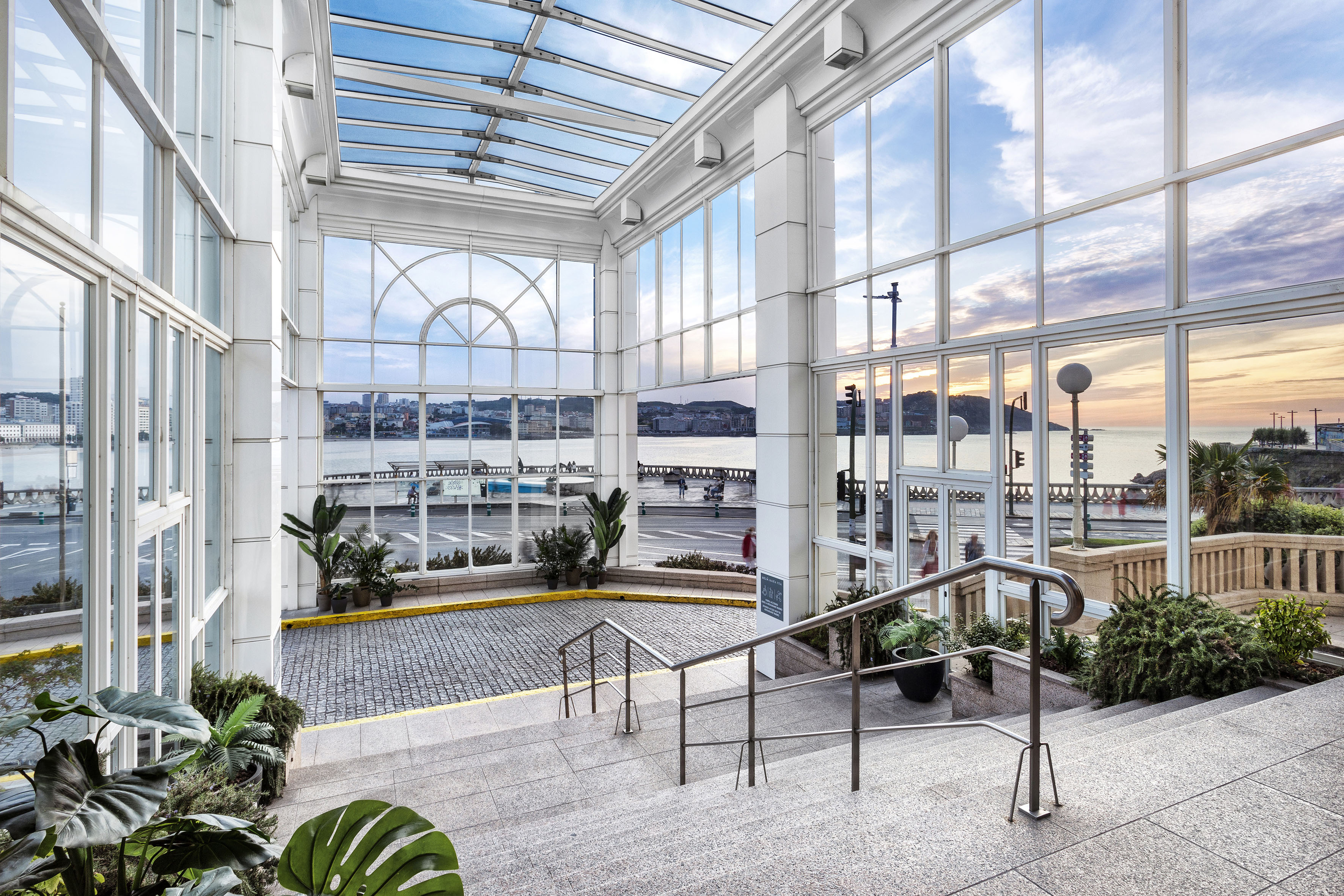 Modern glass atrium with panoramic bay view, stairs, and sunset sky.