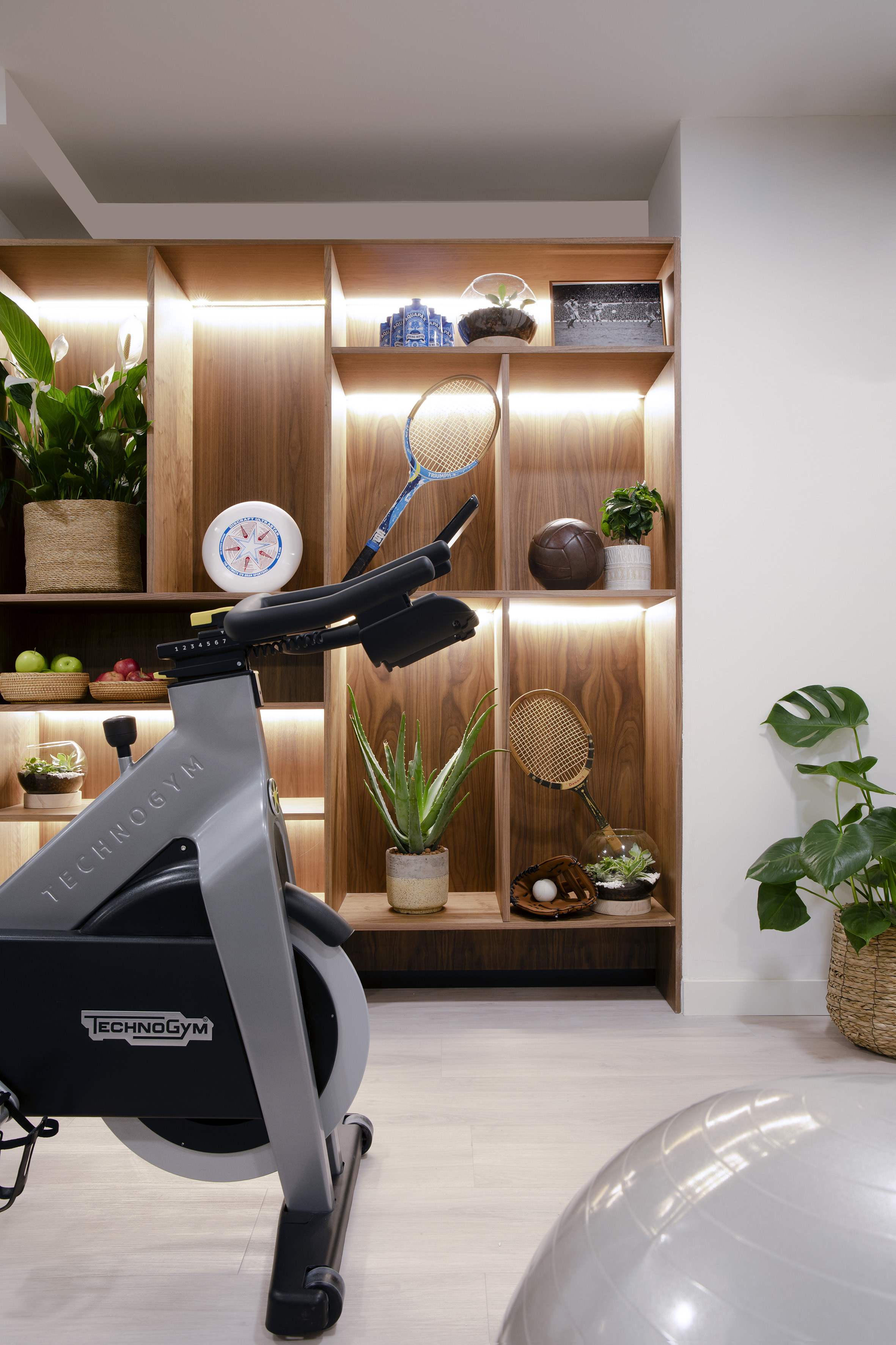Stylish home gym with Technogym bike, plants, and vintage sports equipment on wooden shelves.