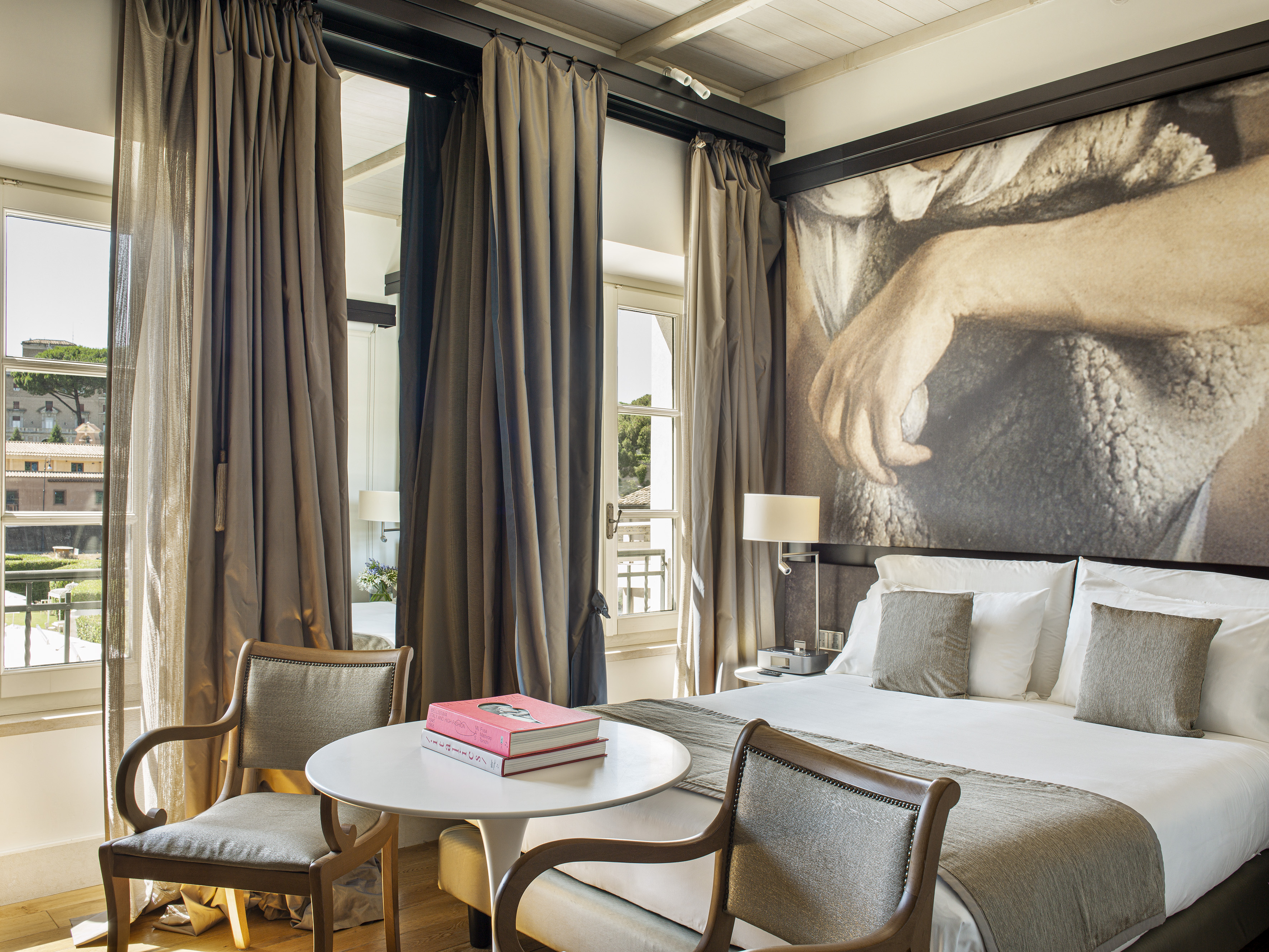 Luxurious hotel bedroom with large bed, classical art mural, seating area, and city view.