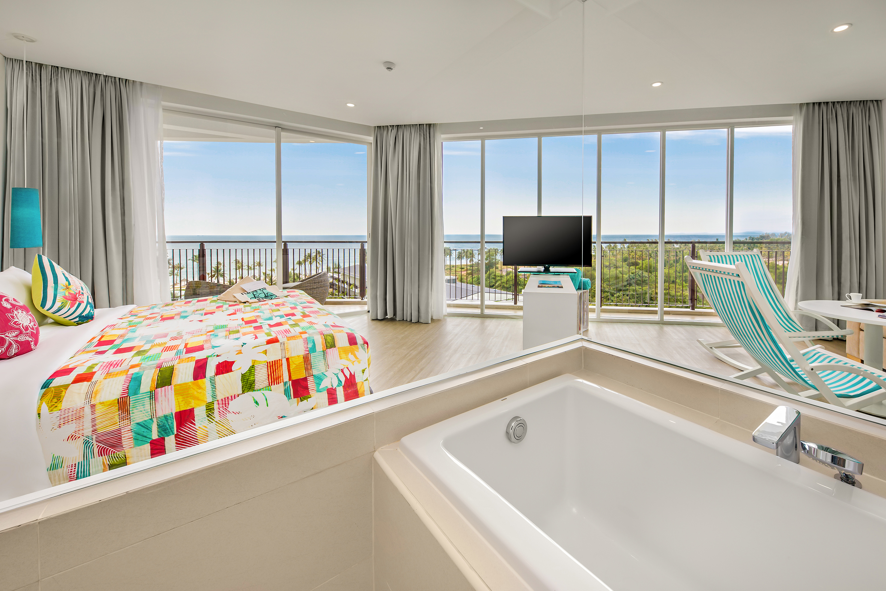 Ocean view hotel suite with colorful bed, balcony, and in-room bathtub.