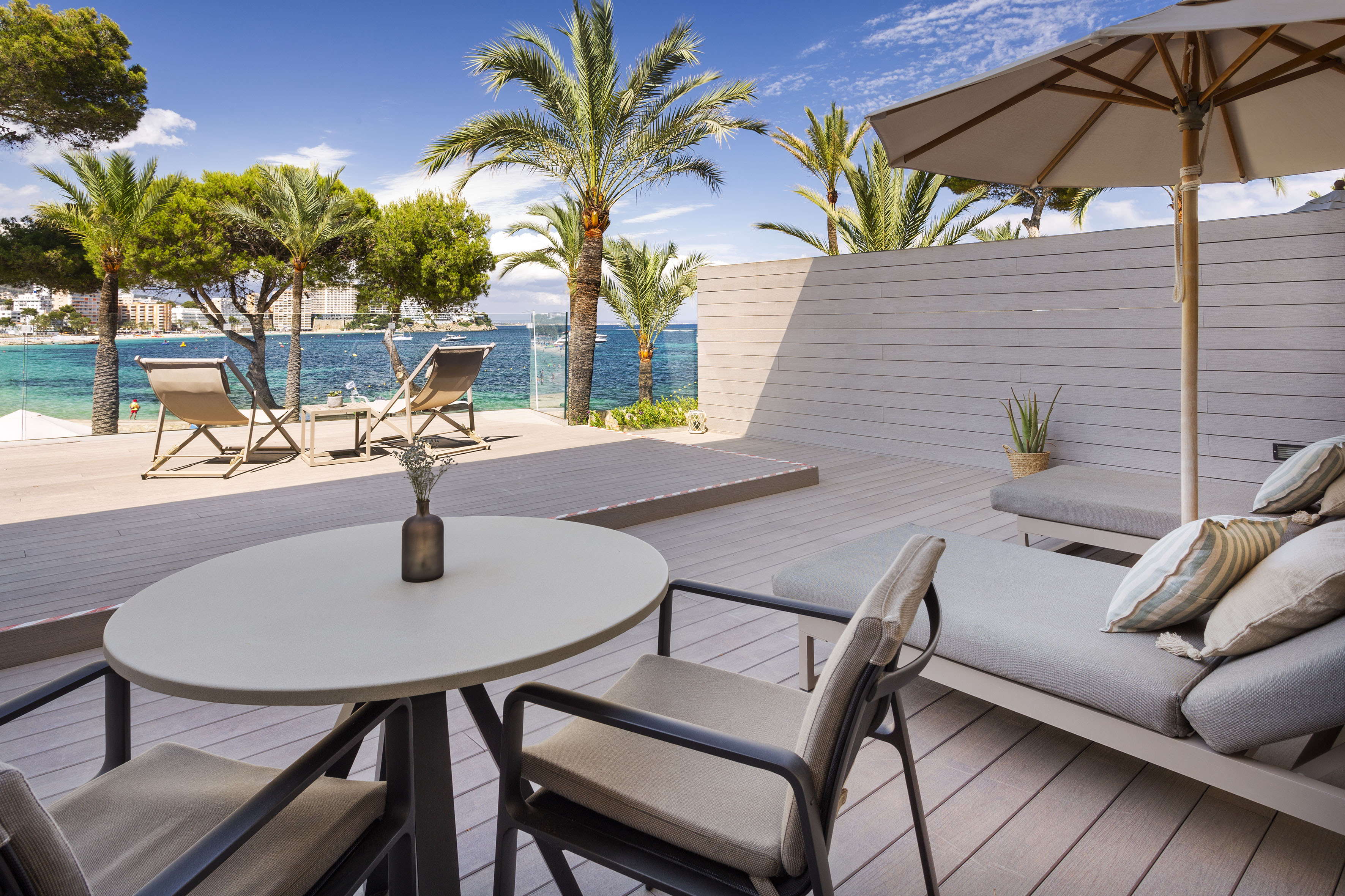 Beach resort patio with modern lounge furniture, palm trees, and blue sea view.