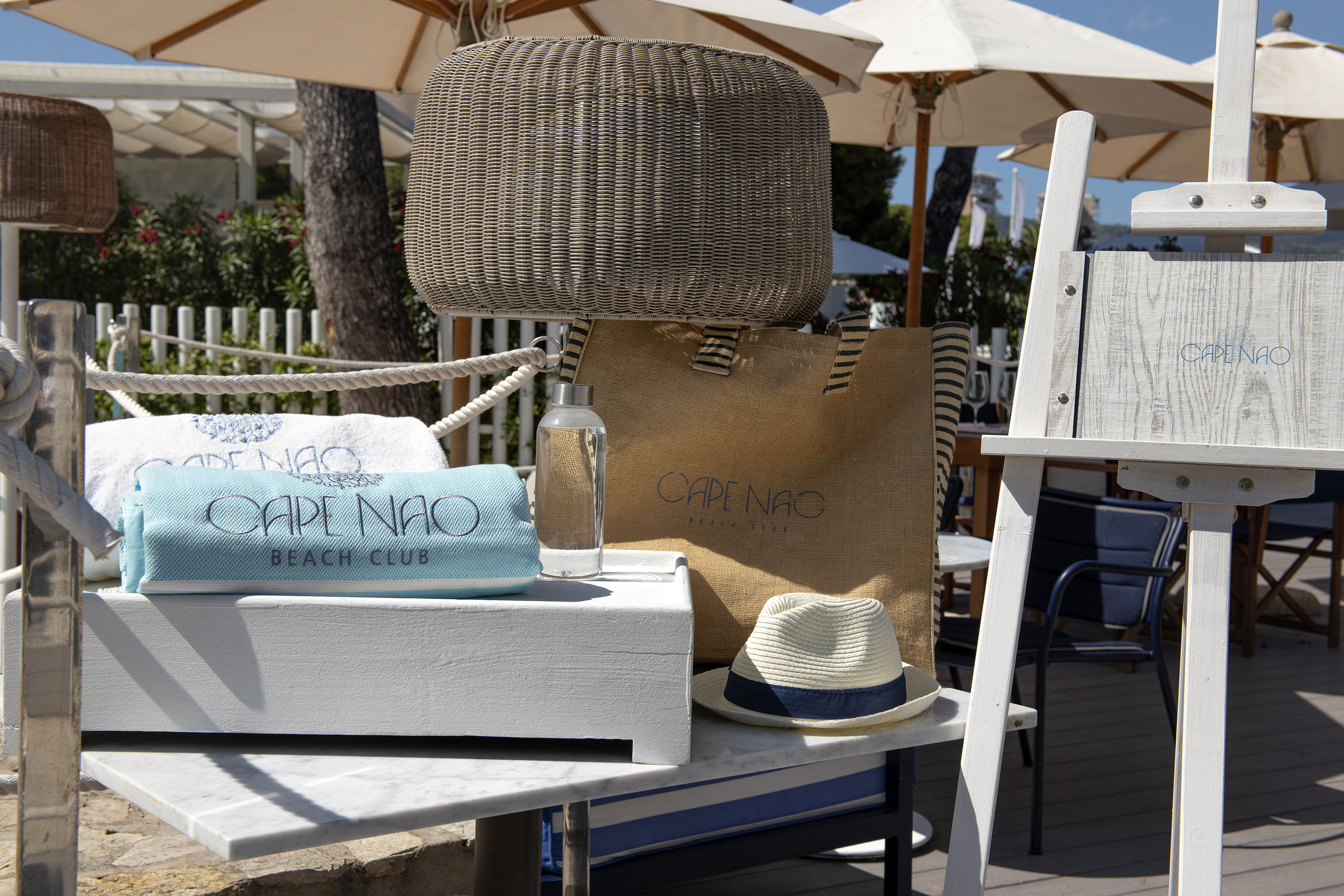 Cape Nao Beach Club gear: towels, tote, hat, water bottle, wicker basket.