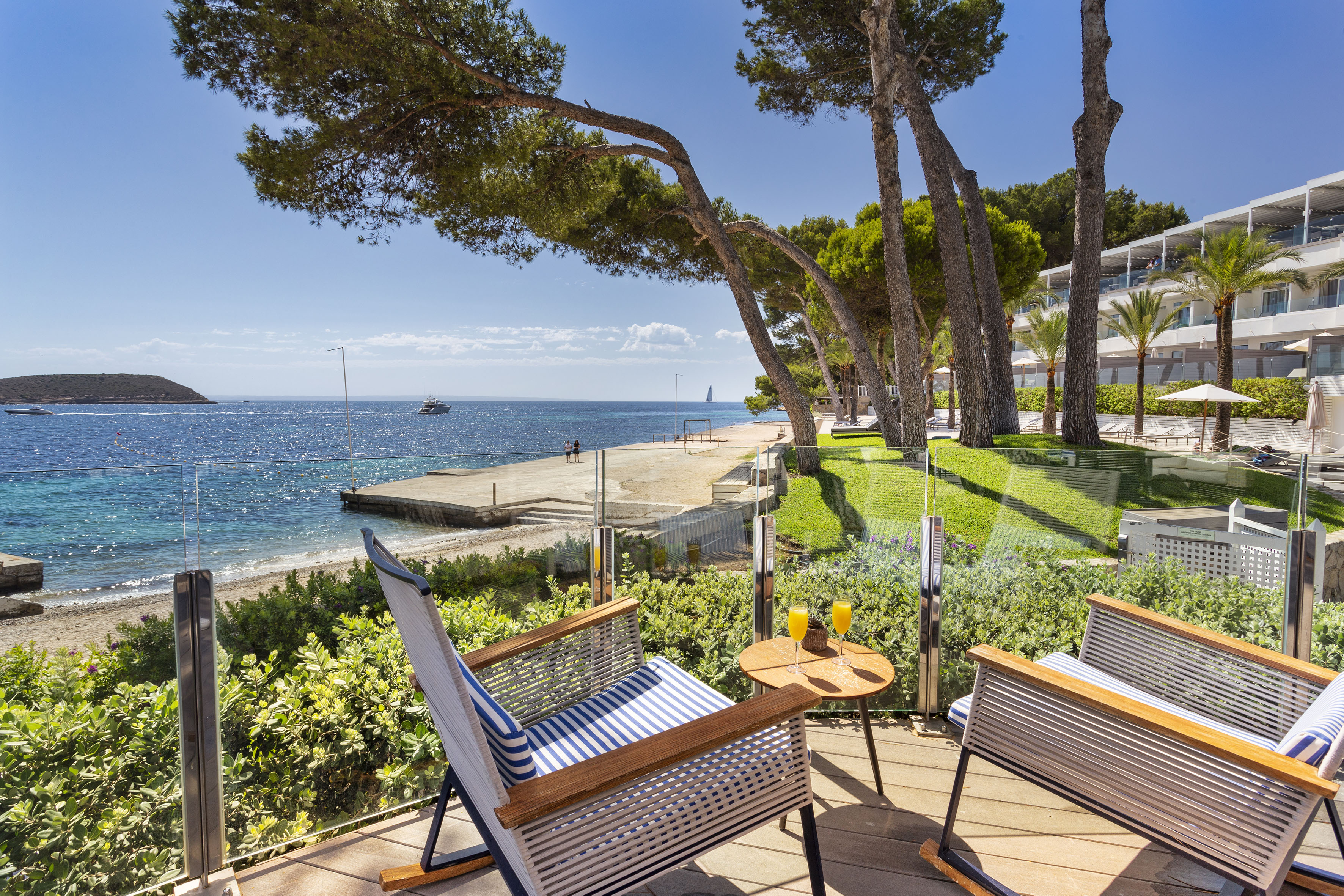 Hotel balcony with chairs, cocktails, ocean view, sandy beach, and pine trees.