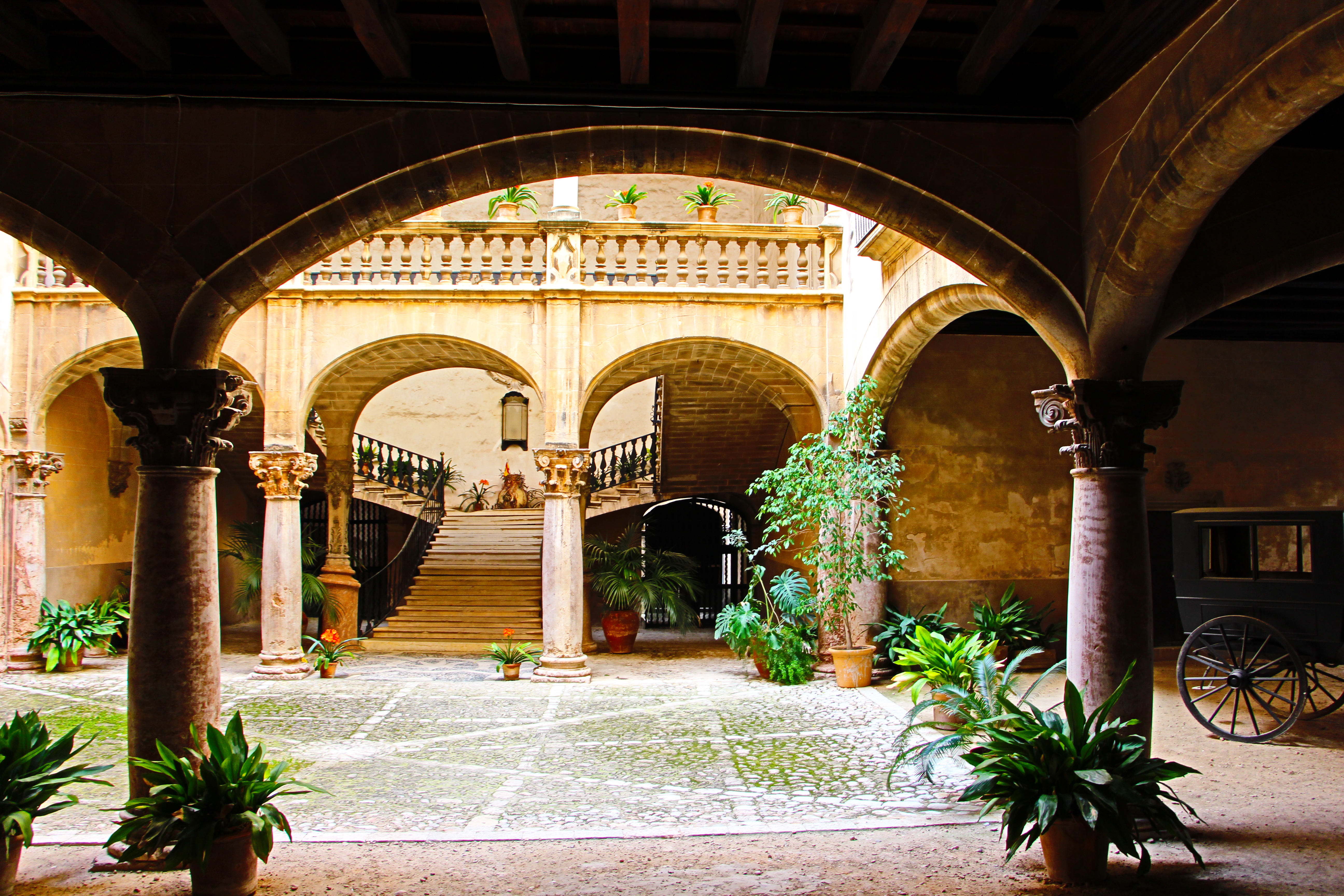 Historic courtyard with arched columns, stairs, potted plants, and vintage carriage.