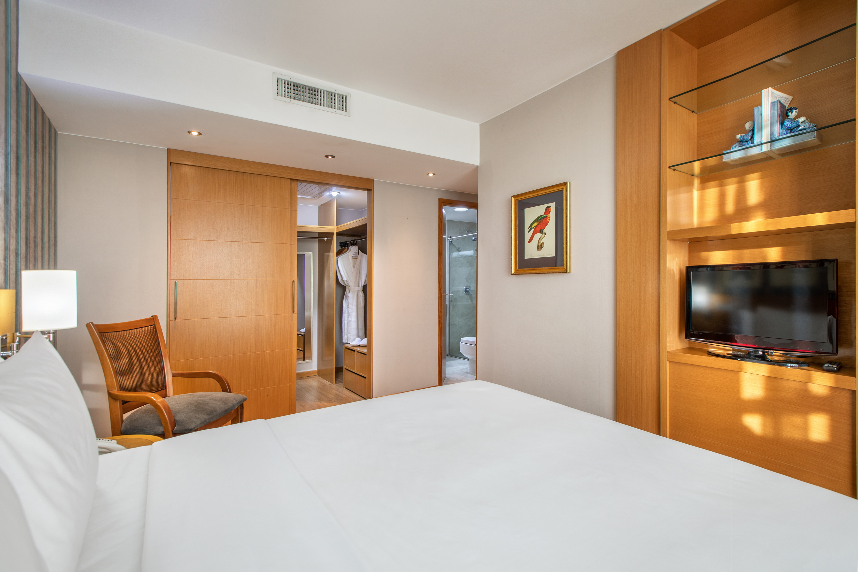 Modern hotel bedroom with white bed, wooden wardrobe, TV, and bathroom.