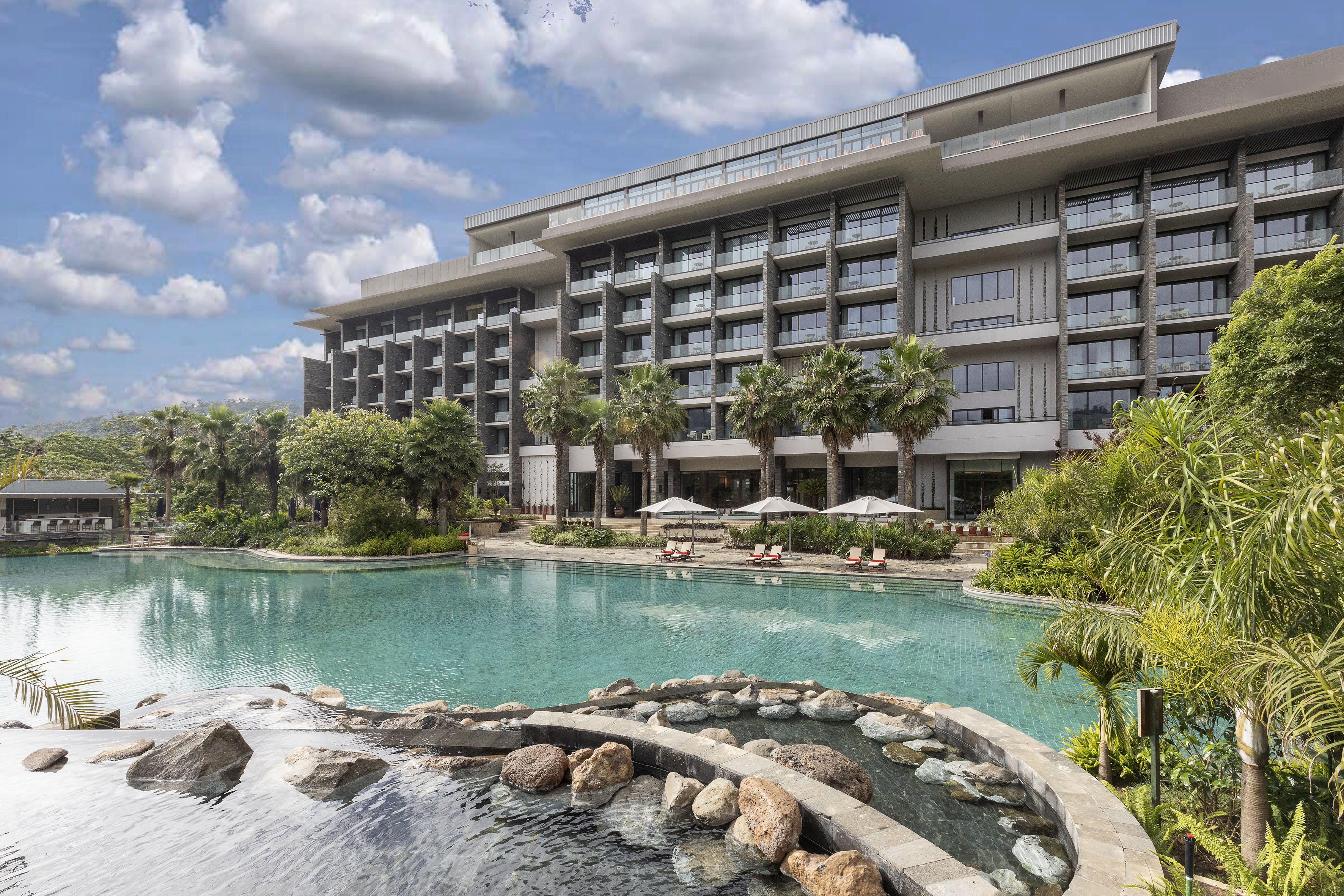 Modern luxury hotel with large swimming pool, palm trees, and balconies.