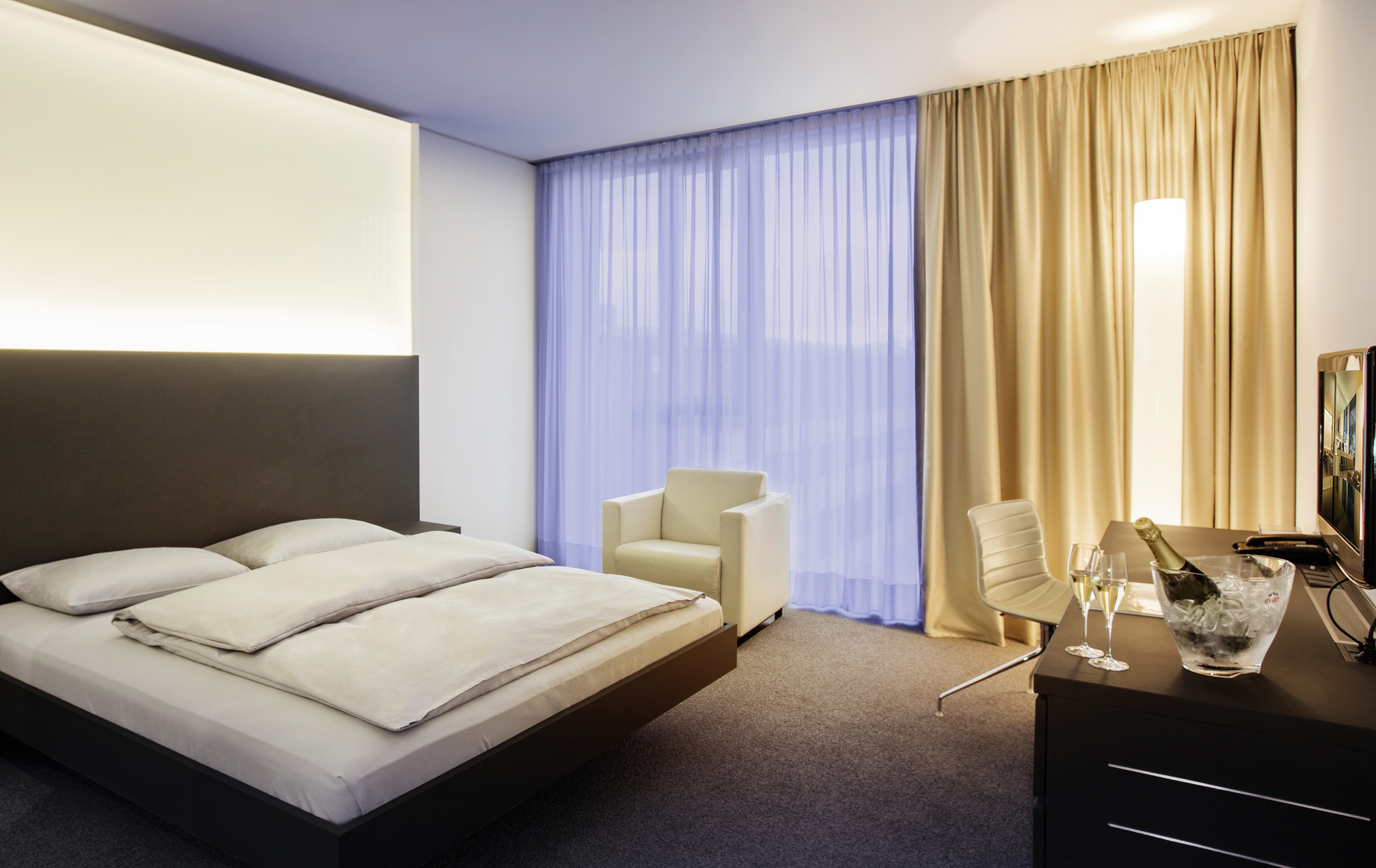 Modern hotel room with bed, glowing headboard, desk, champagne, sheer curtains.