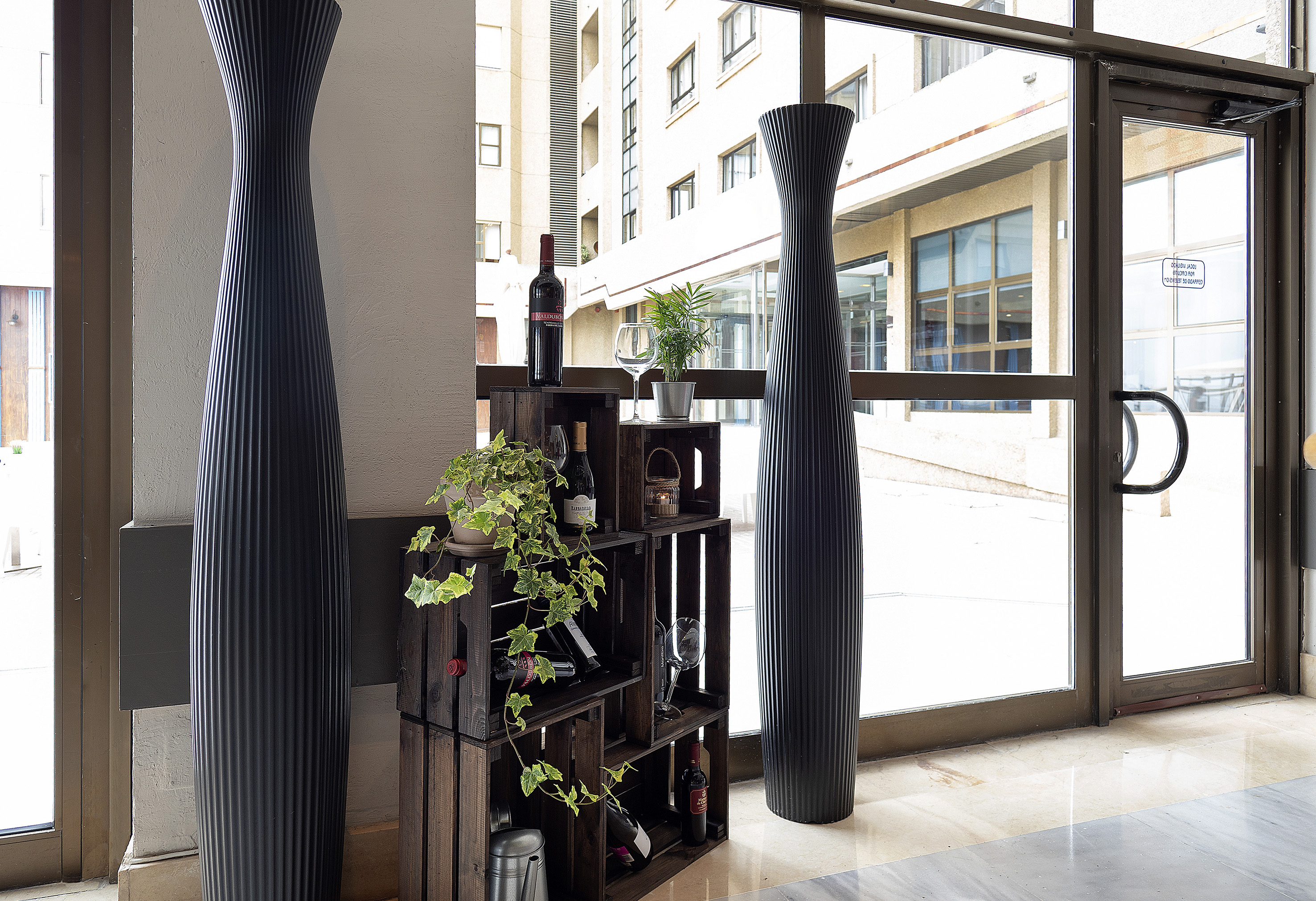 Lobby with tall fluted vases, wine display on crates, plants, and glass entrance.