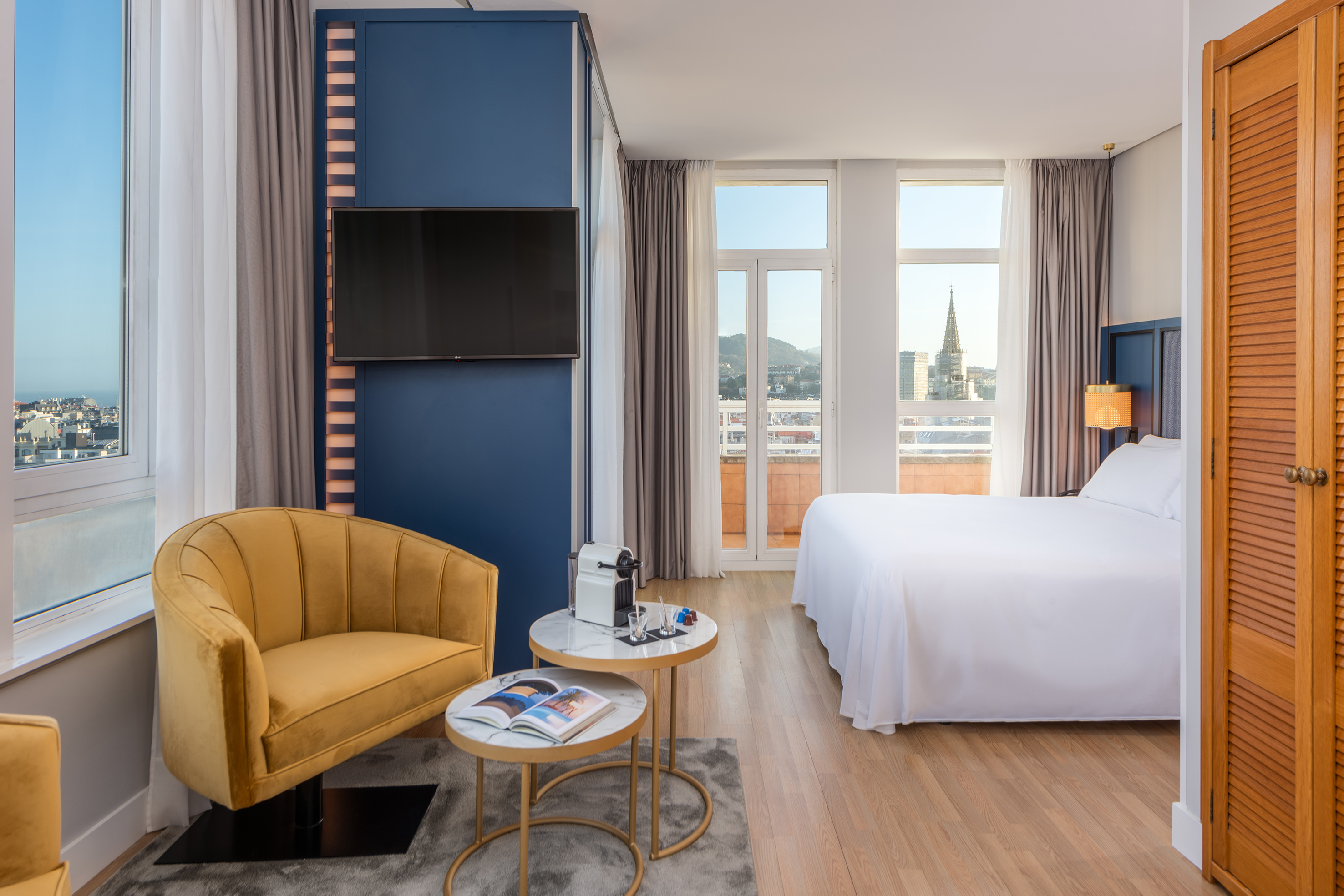 Modern hotel room with city view, yellow velvet armchair, bed, and TV.