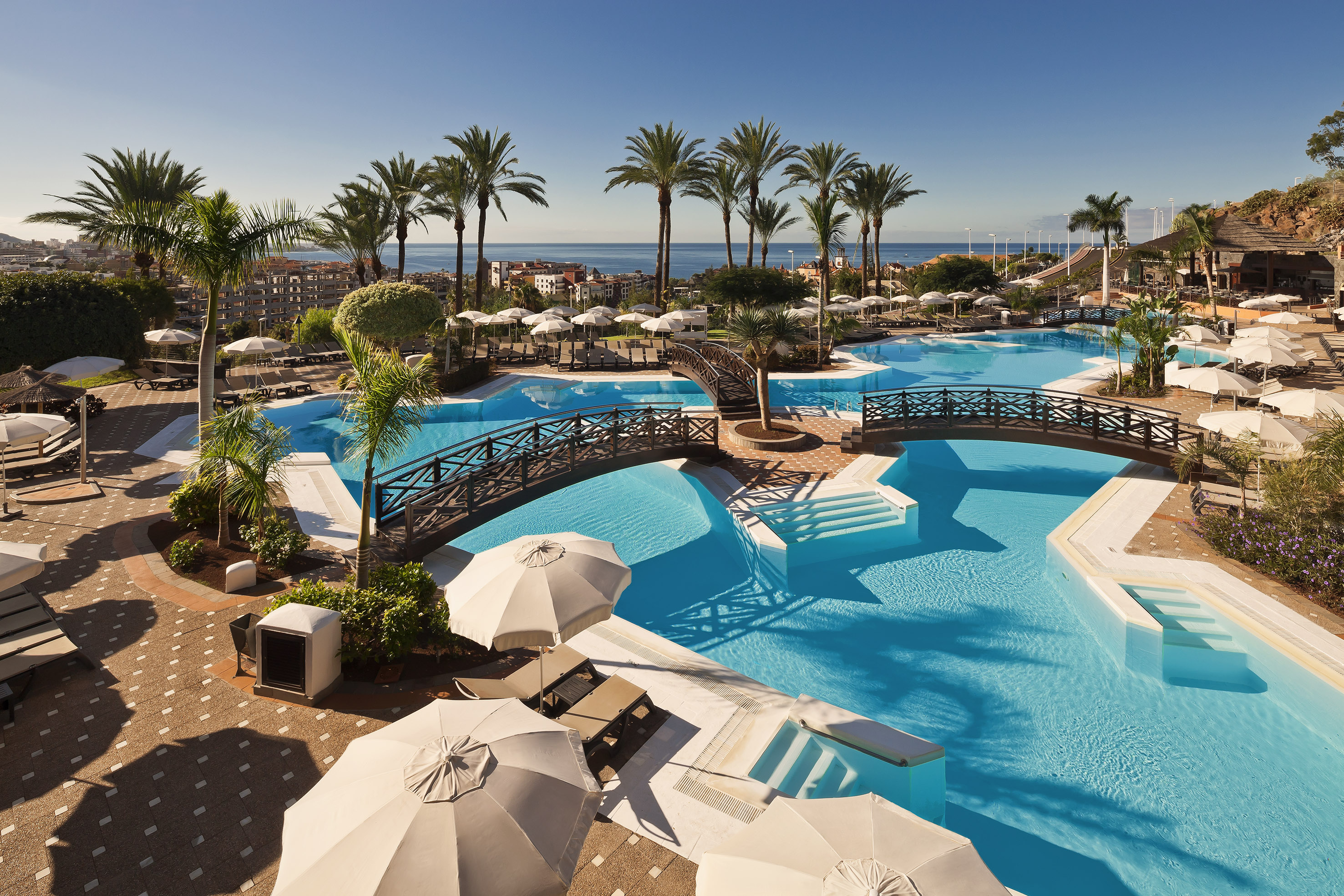 Tropical resort pools with bridges, palm trees, sun loungers, and ocean.