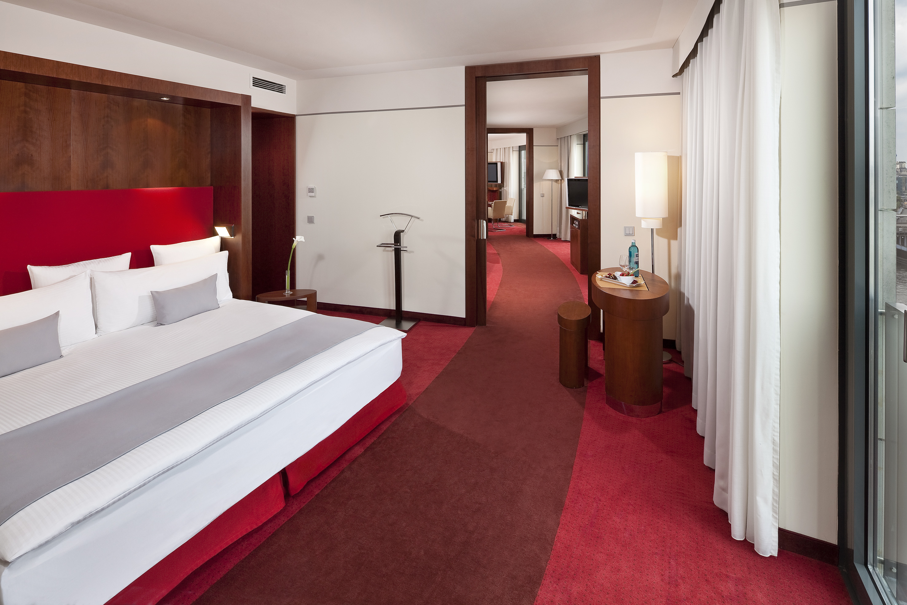 Modern hotel suite with red bed, carpet, and connecting living space.