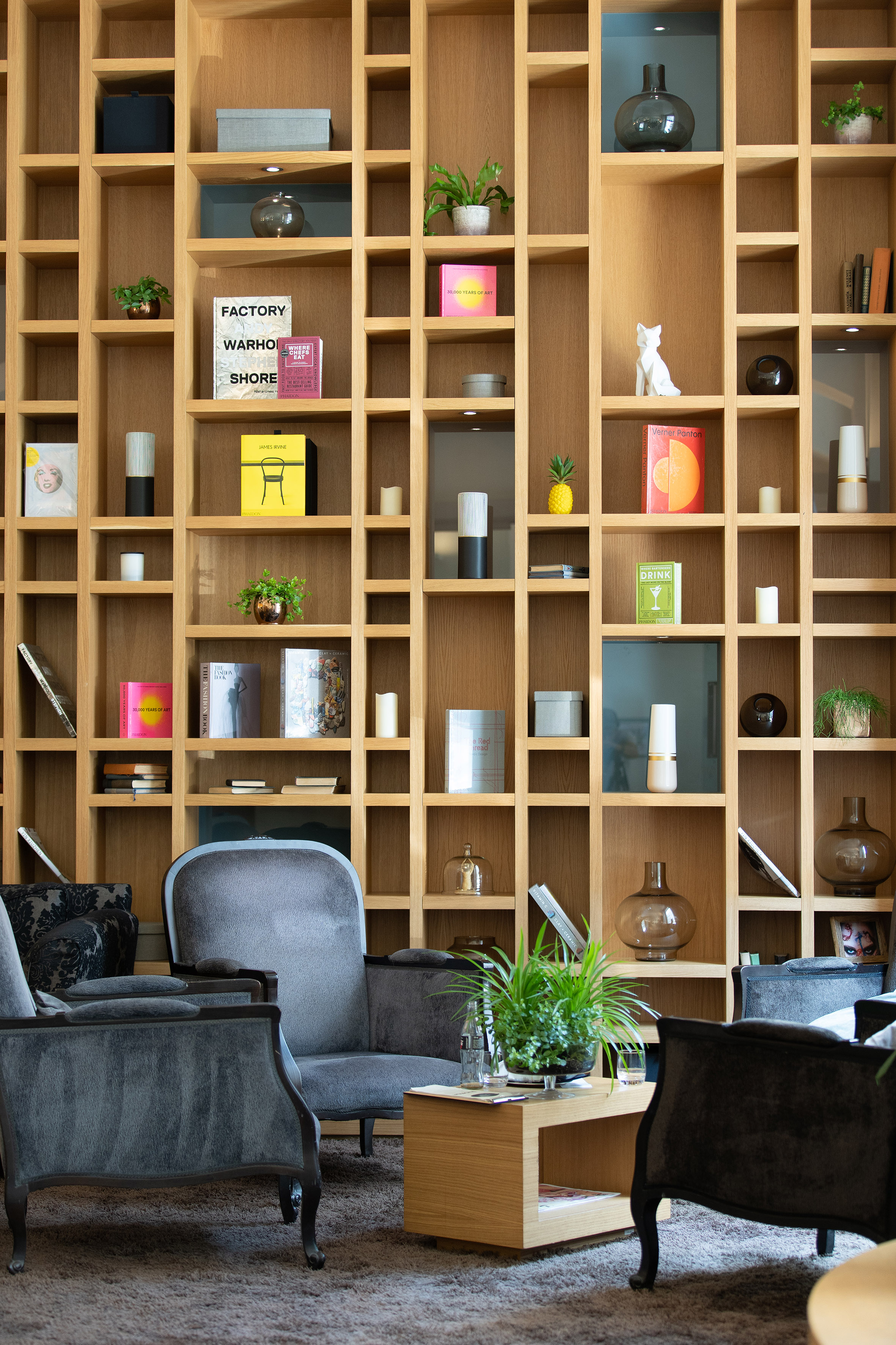 Wooden bookshelf wall with books, plants, and decor, modern lounge seating.
