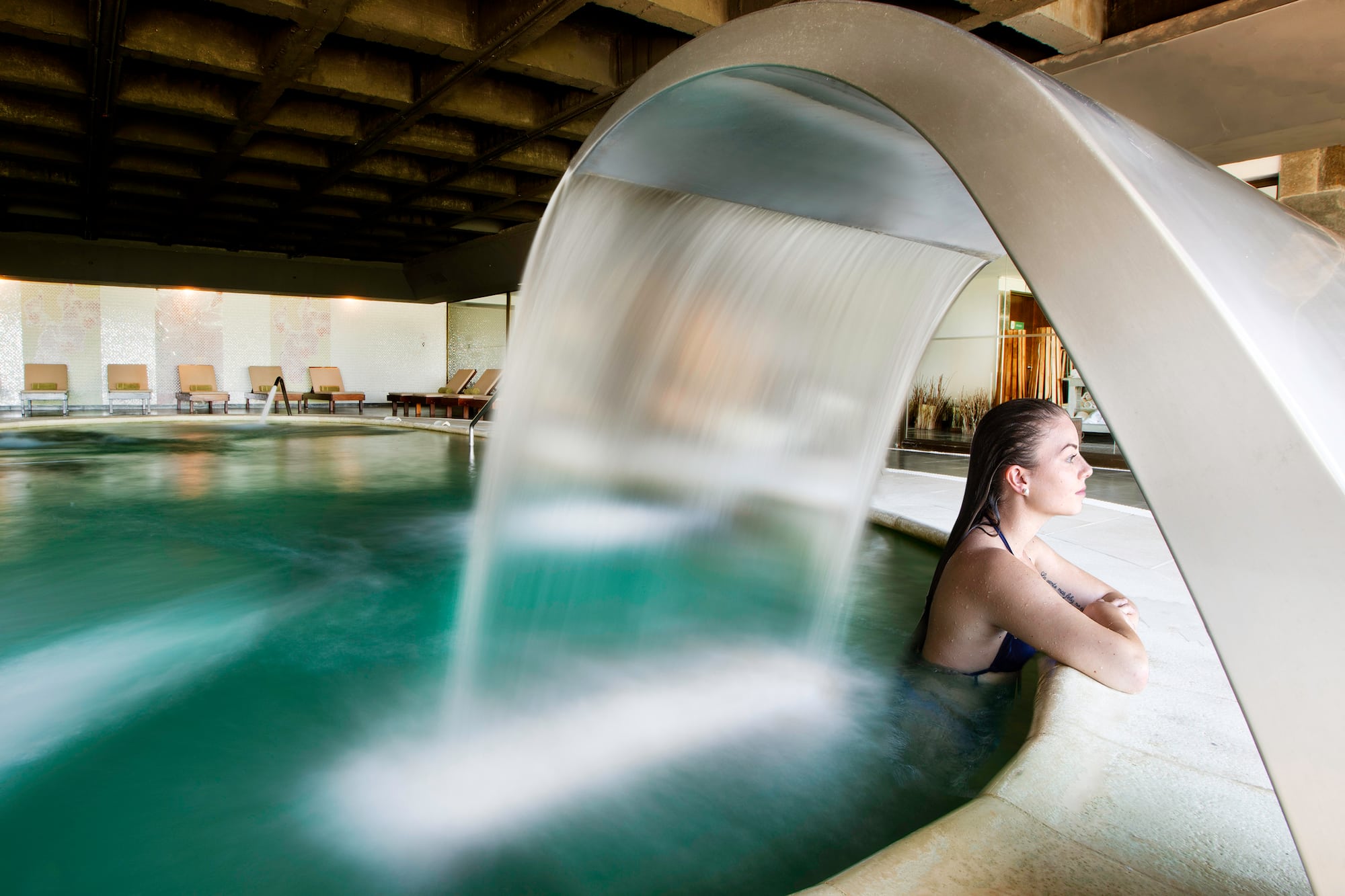 Woman relaxing in indoor spa pool with hydrotherapy waterfall.