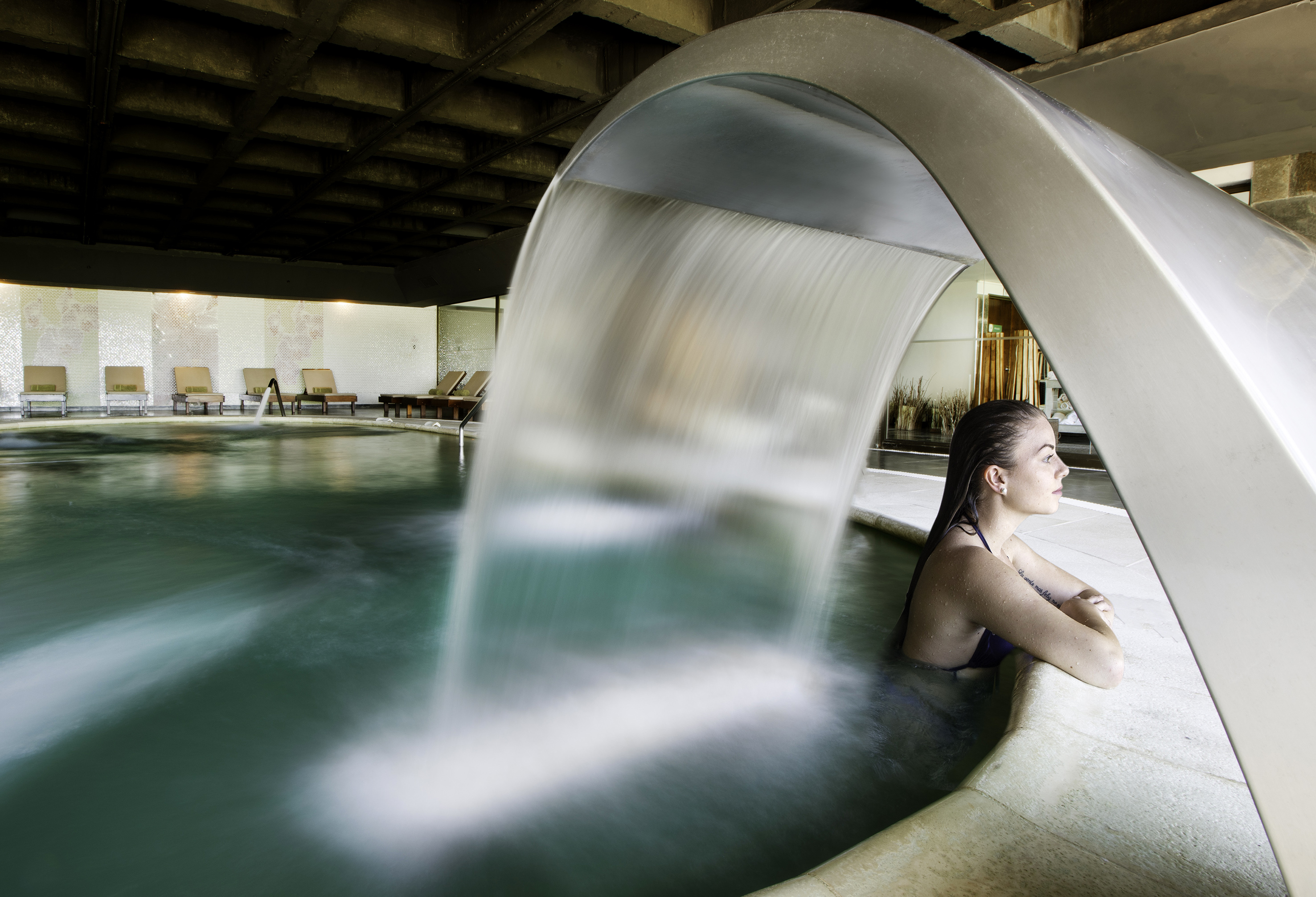 Woman relaxing in indoor spa pool with hydrotherapy waterfall.