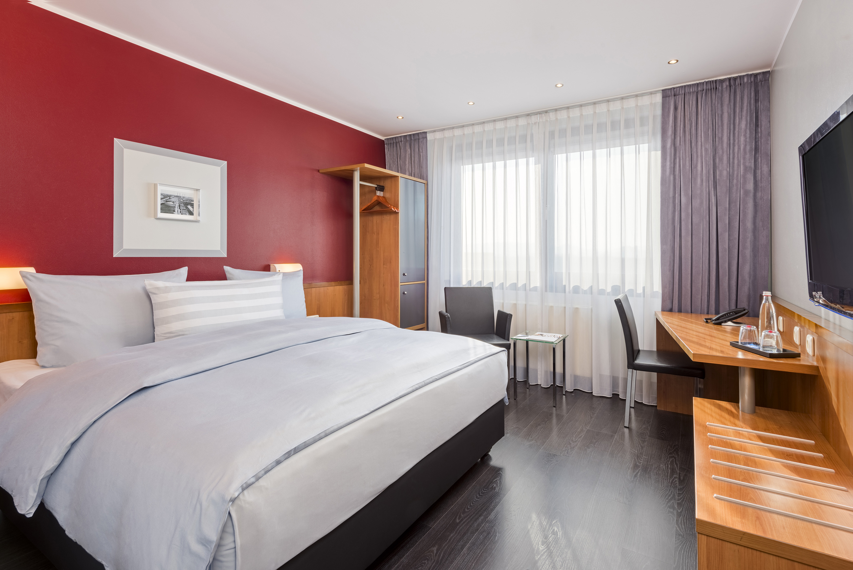 Modern hotel bedroom with red accent wall, bed, desk, and window.