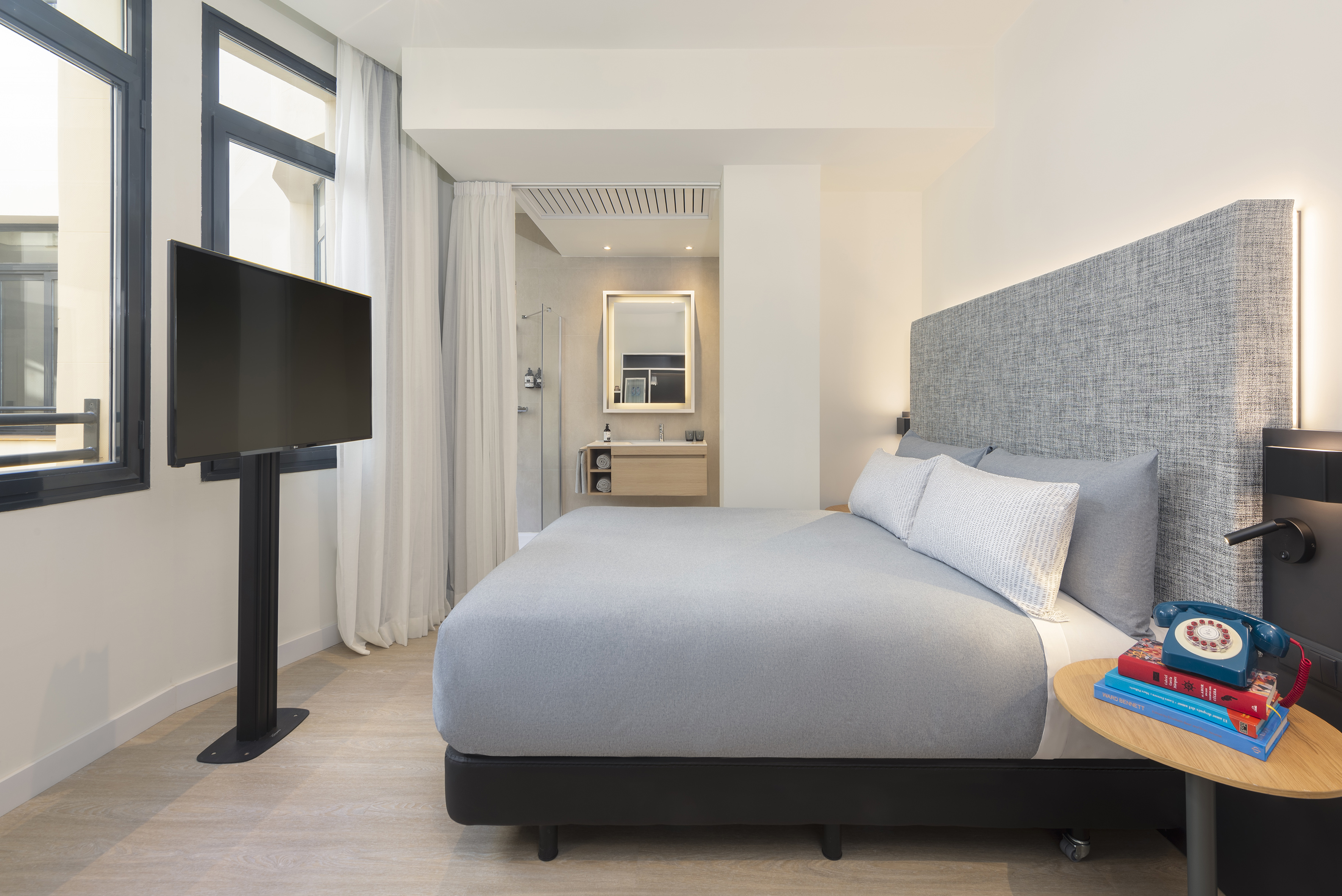 Modern hotel room with grey bed, wall TV, and open-plan bathroom.