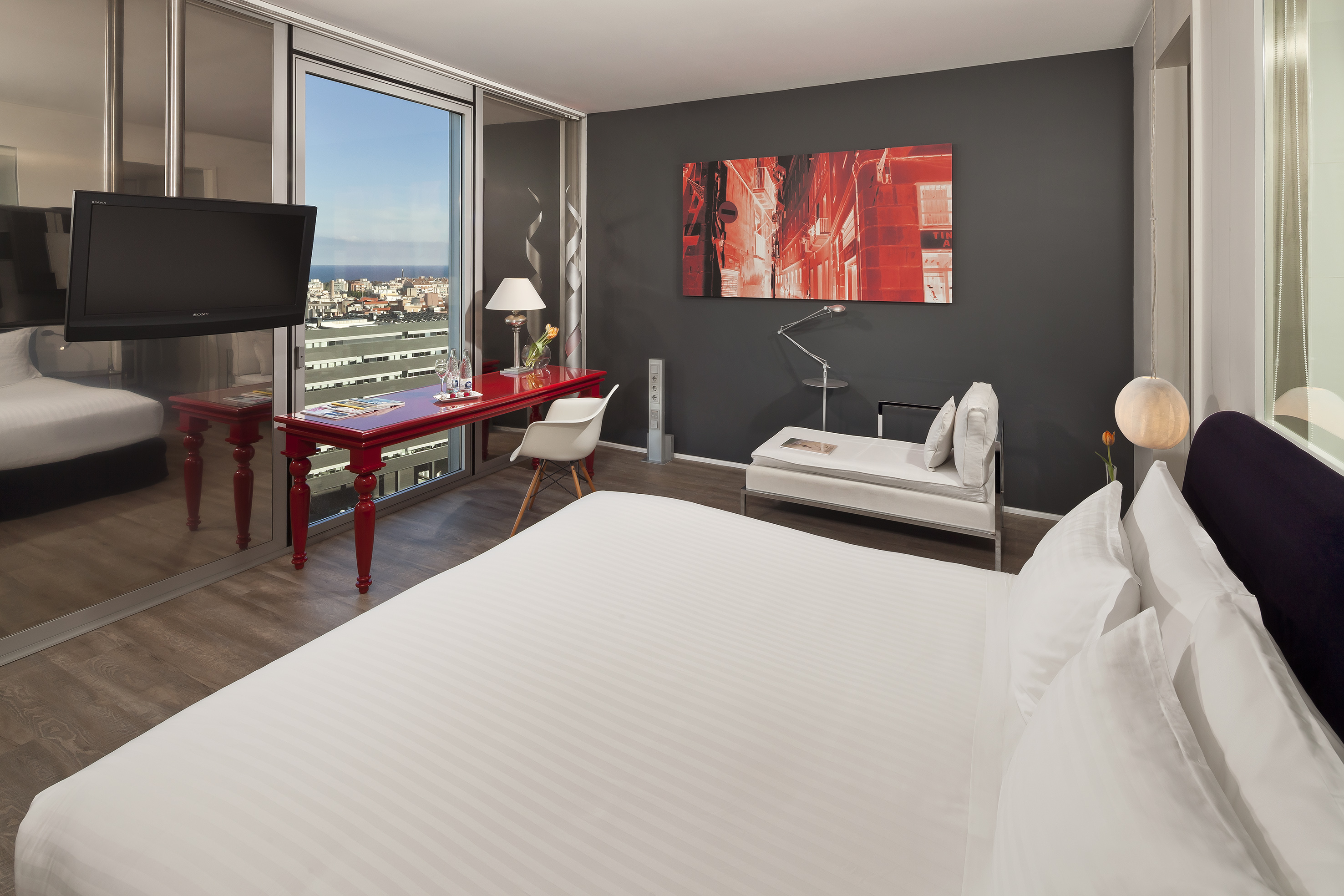 Hotel room with panoramic city & sea view, white bed, red desk, modern design.